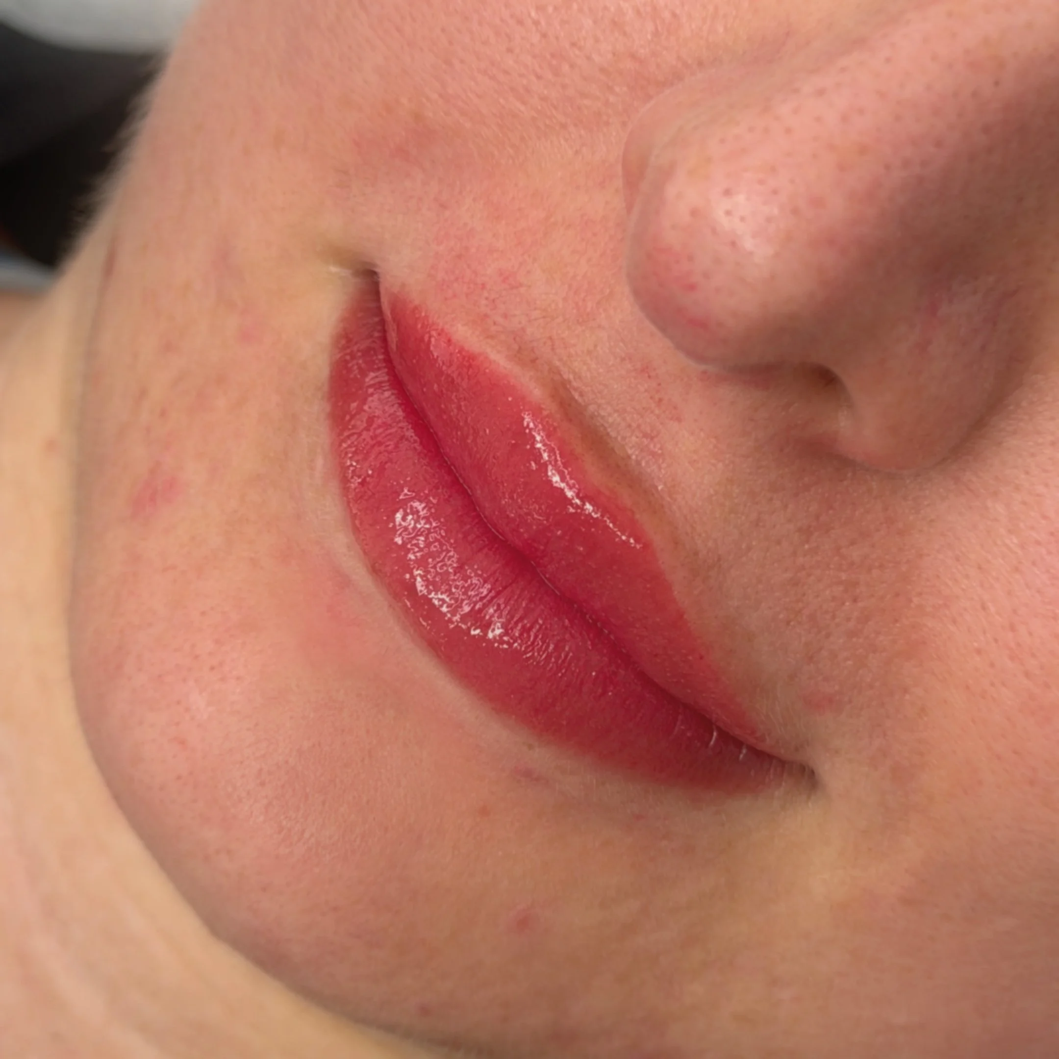 Close-up of fresh lip blush tattoo with gloss, partially visible nose and cheek, and smooth skin.