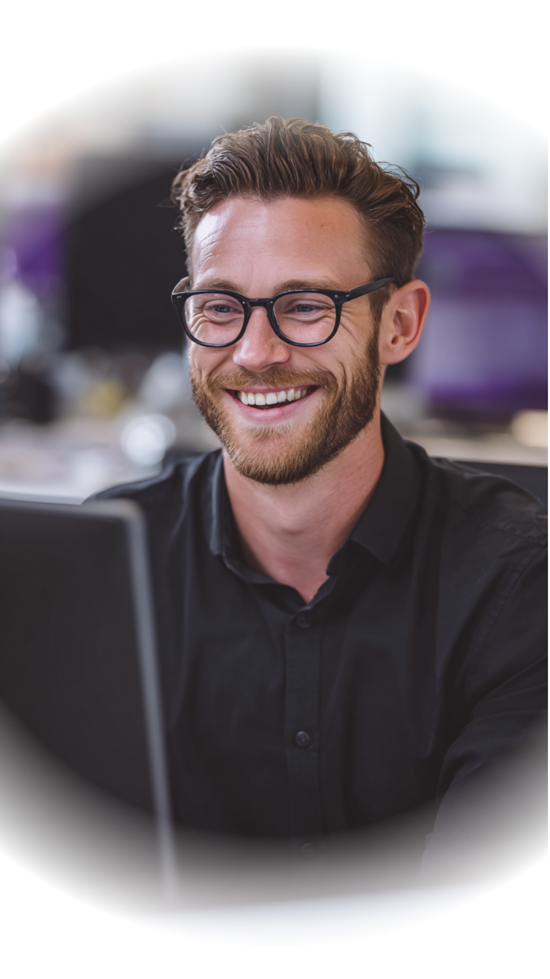 A young man with glasses and a beard smiling while working at a desk in an office setting