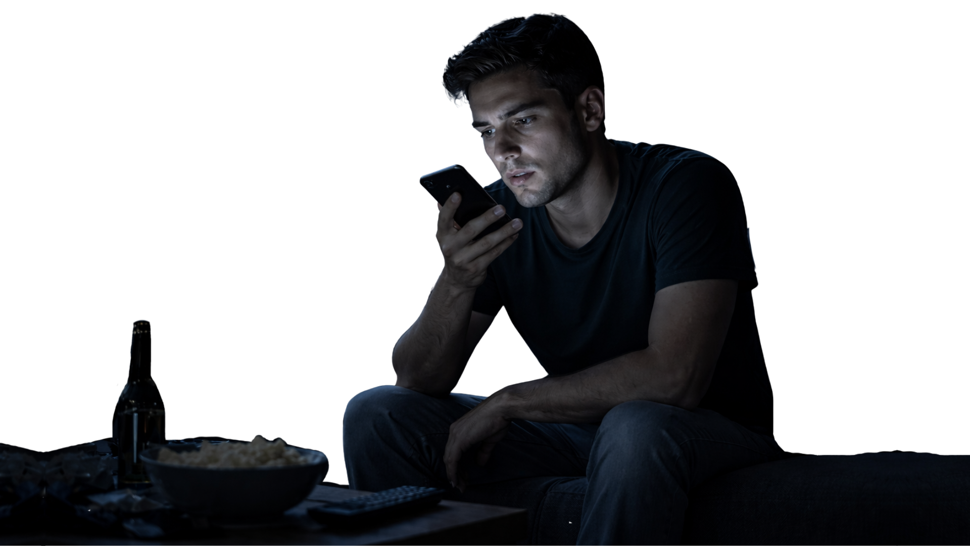 A man sitting on a couch in a dark room looking at his phone with a bowl of popcorn, a remote control, an open beer bottle, and snack wrappers on the table in front of him.
