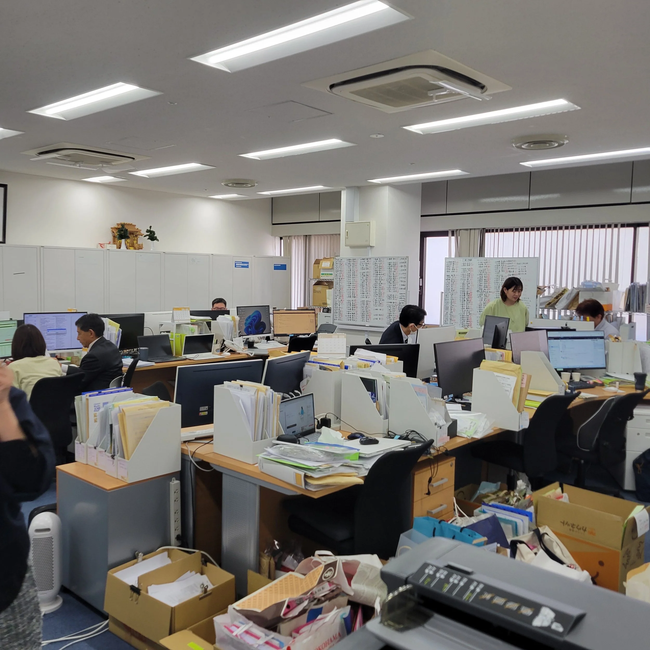 オフィスのデスクや棚に書類や紙類が散乱しており、複数のコンピューターと社員が作業している様子が映っている