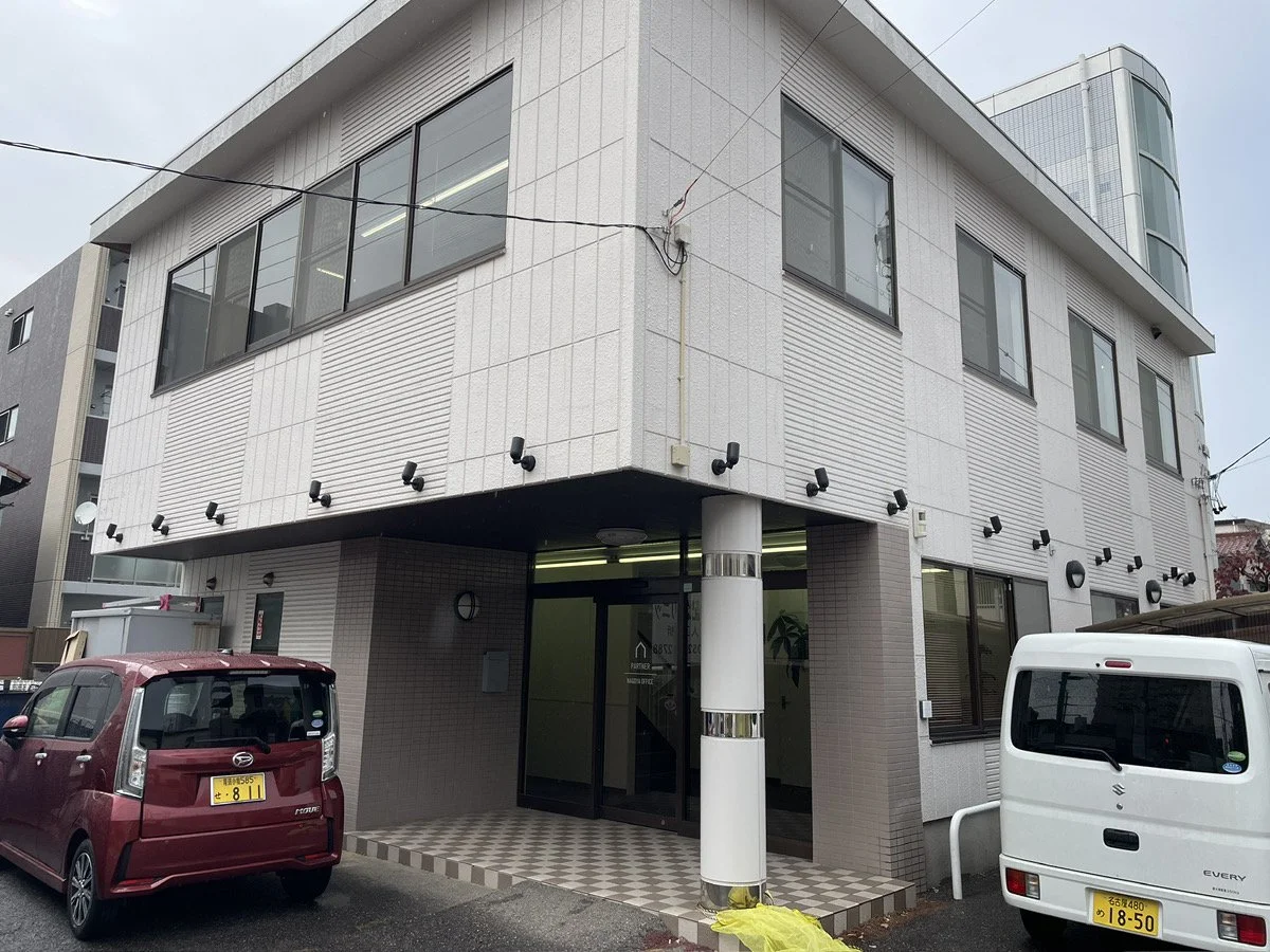 白い三階建ての建物、駐車場に赤と白の車が停まっている。建物の入り口にはガラス扉と壁に照明とスピーカーが設置されている。
