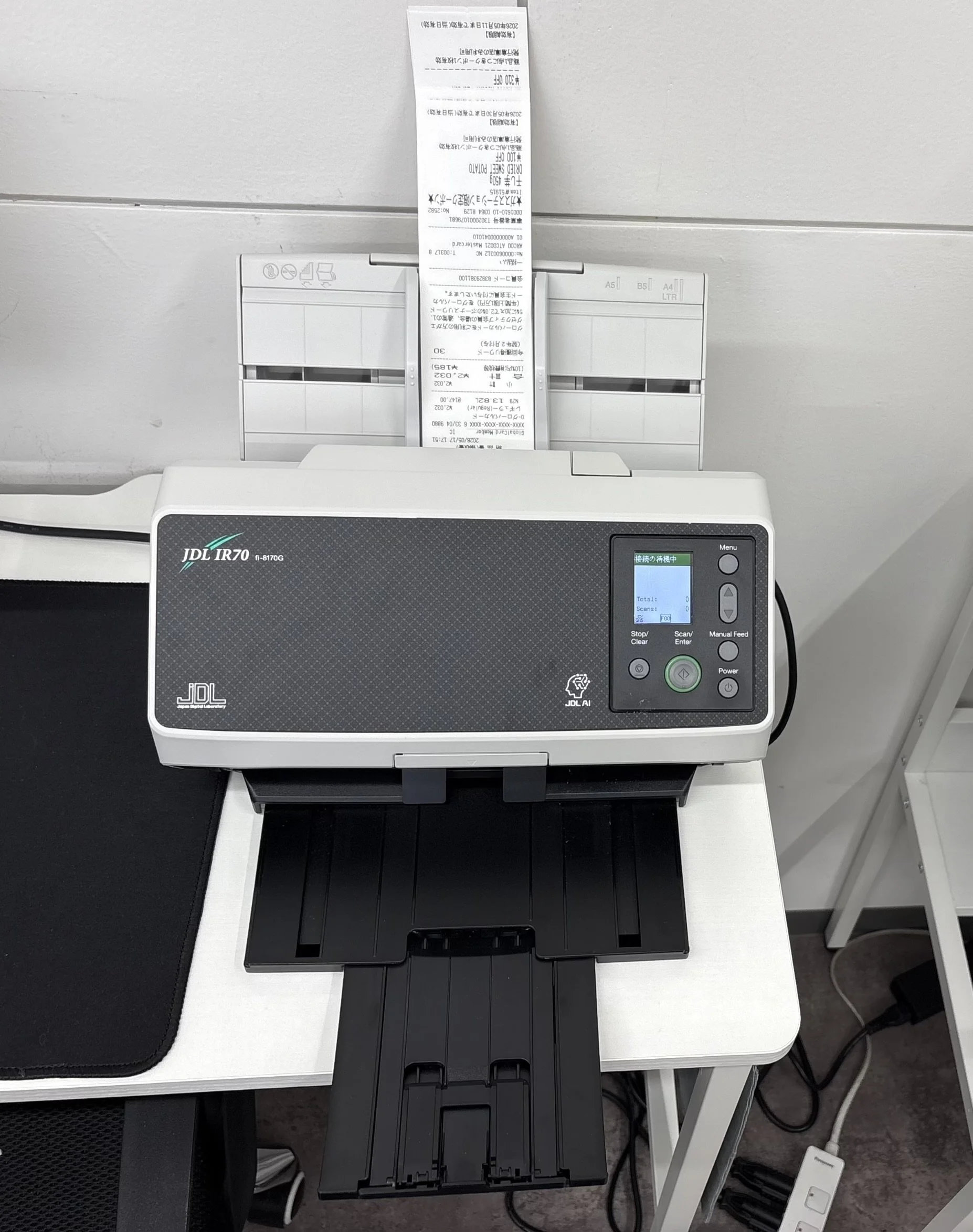 プリンターの上に長い紙のレシートが置かれ、その下に書類や黒い色のトレイが設置された白い机の上の写真