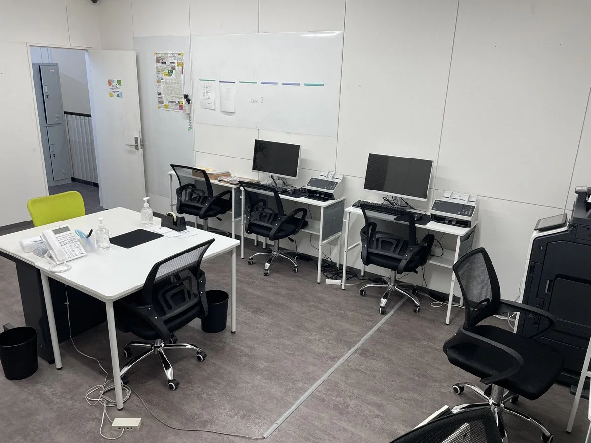 オフィスの会議室または作業スペースに複数のデスクとコンピュータ、電話、椅子が配置されている。