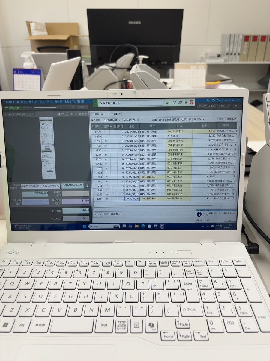 パソコンの画面に会計ソフトのデータと明細が表示されている。背後には複数の書類やオフィス用品が置かれるデスク環境。