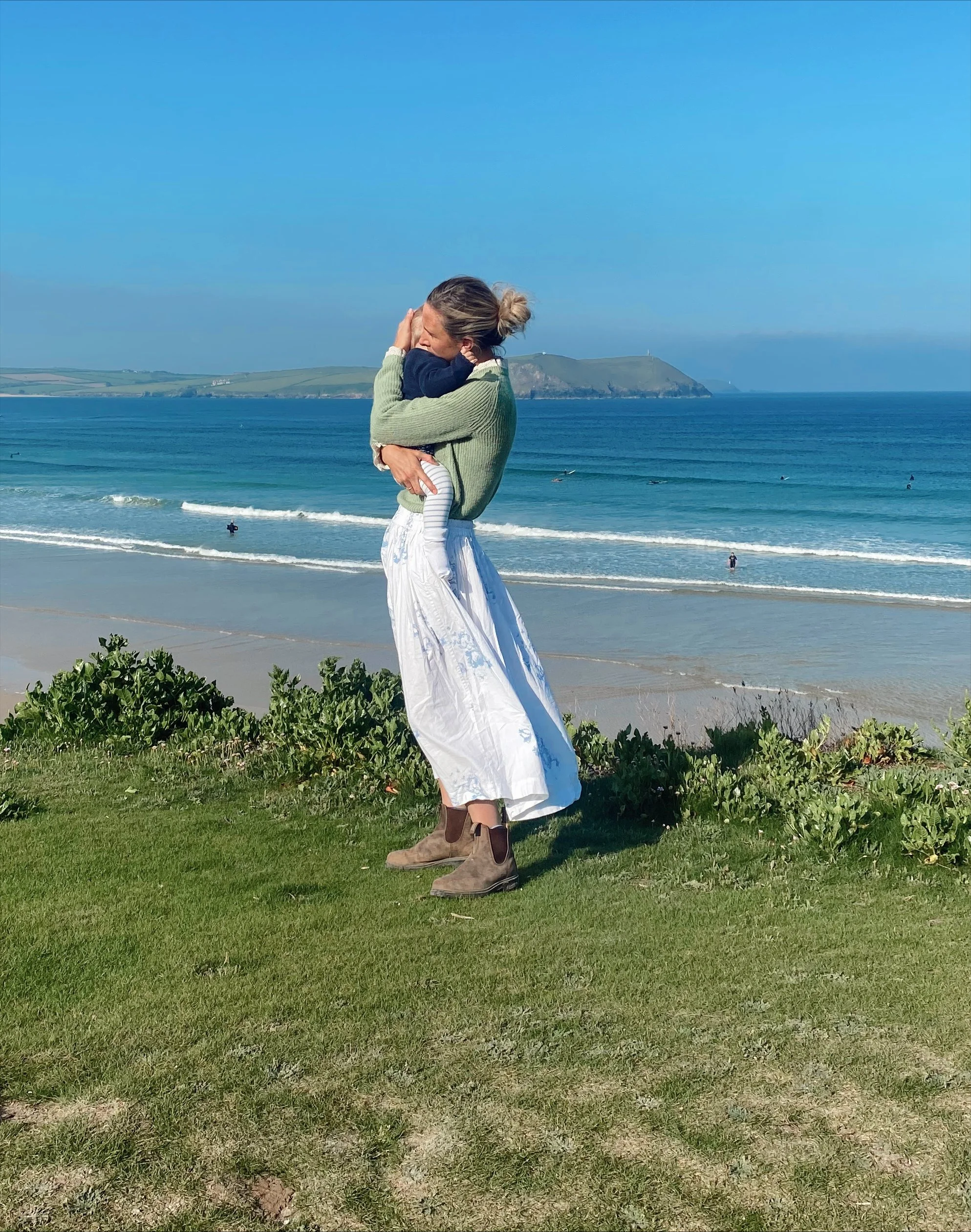 A woman hugging a child on a grassy beach with the ocean and cliffs in the background.