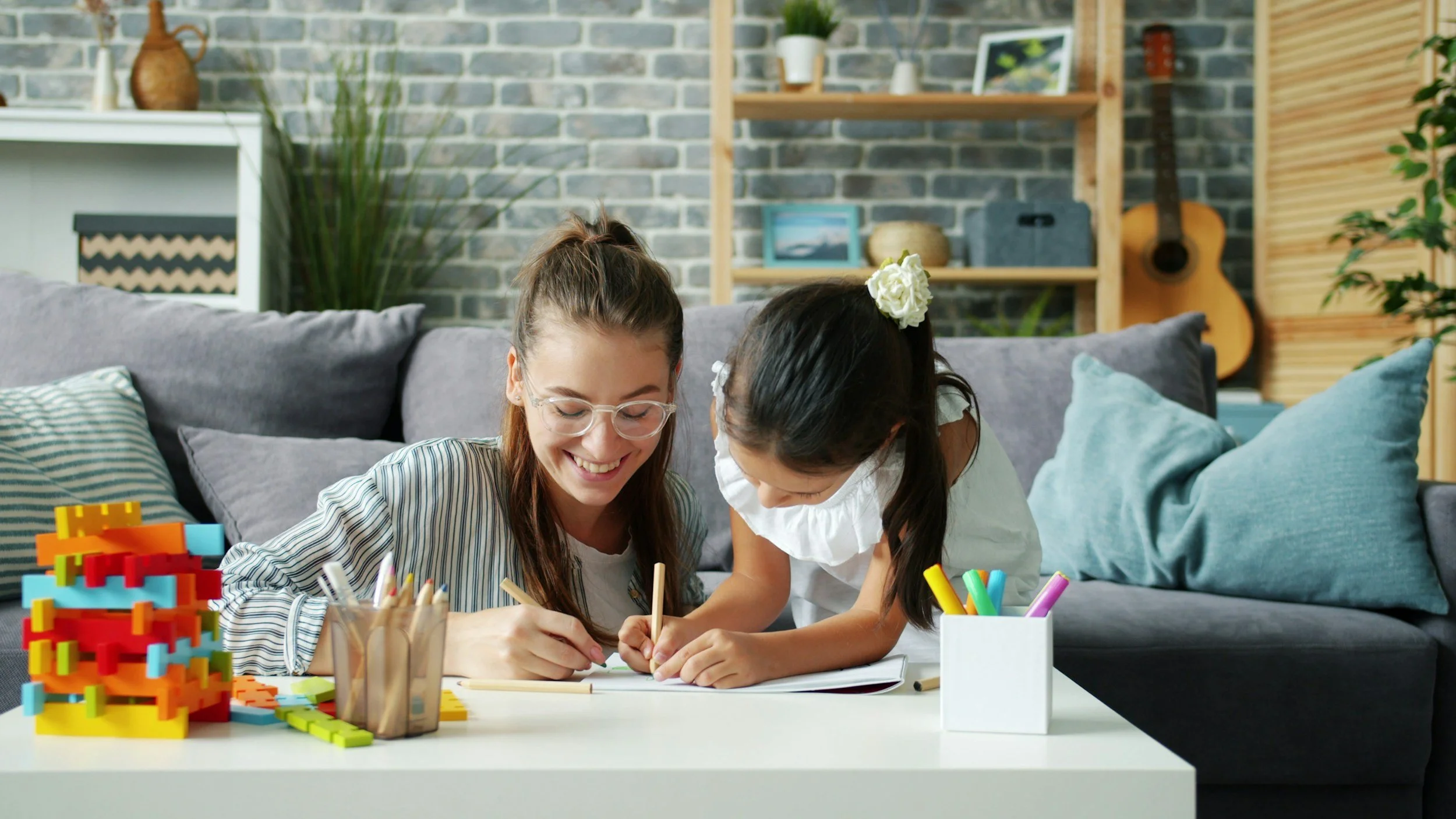 A woman and a young girl are drawing together at a white table in a cozy living room with a gray sofa, pillows, a wooden wall, a guitar, and decorative shelves in the background.