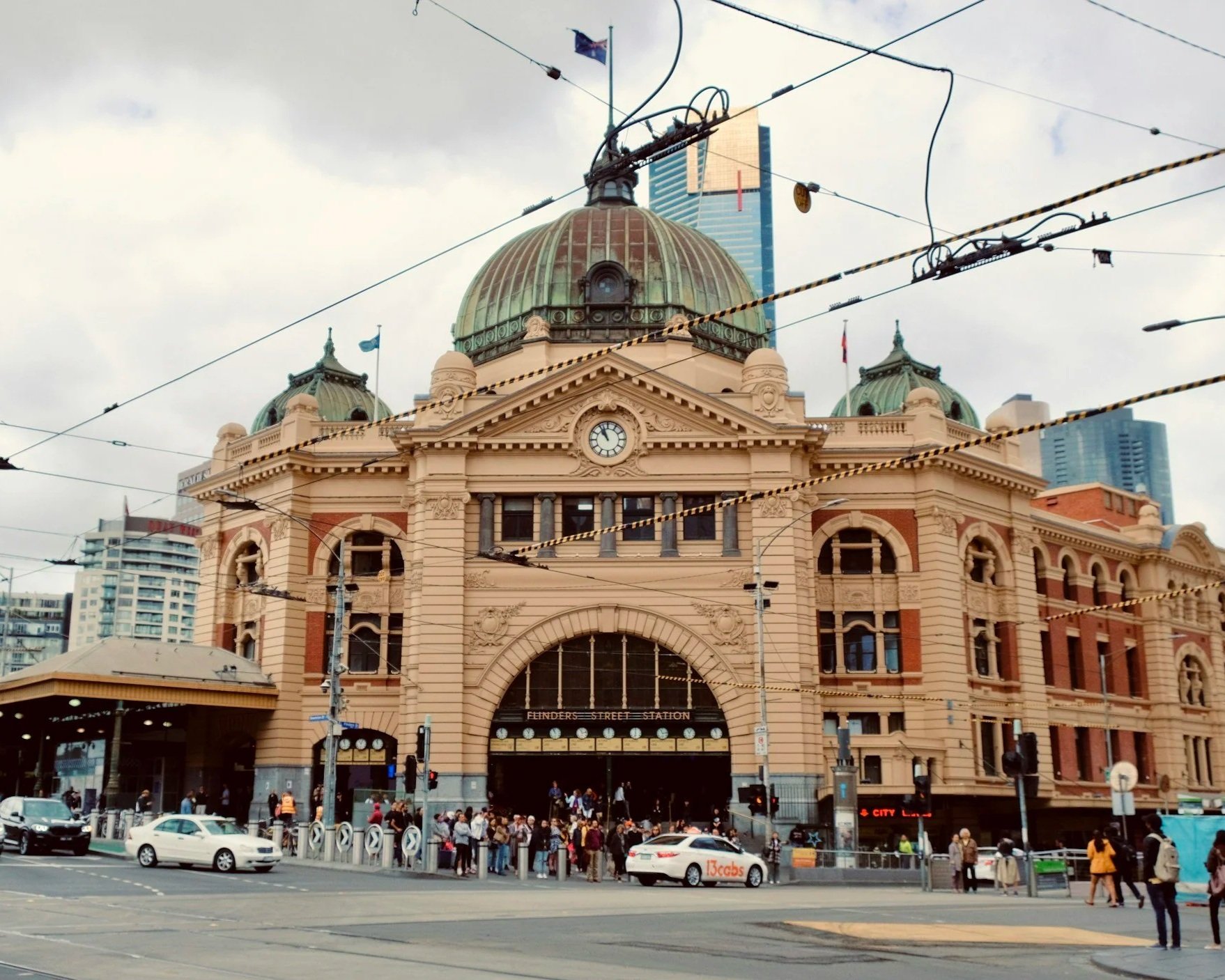 Fitzroy Gardens, historic building with domed roof, clock, and crowd outside, busy city street with cars and pedestrians, overhead tram wires, modern skyscrapers in the background.