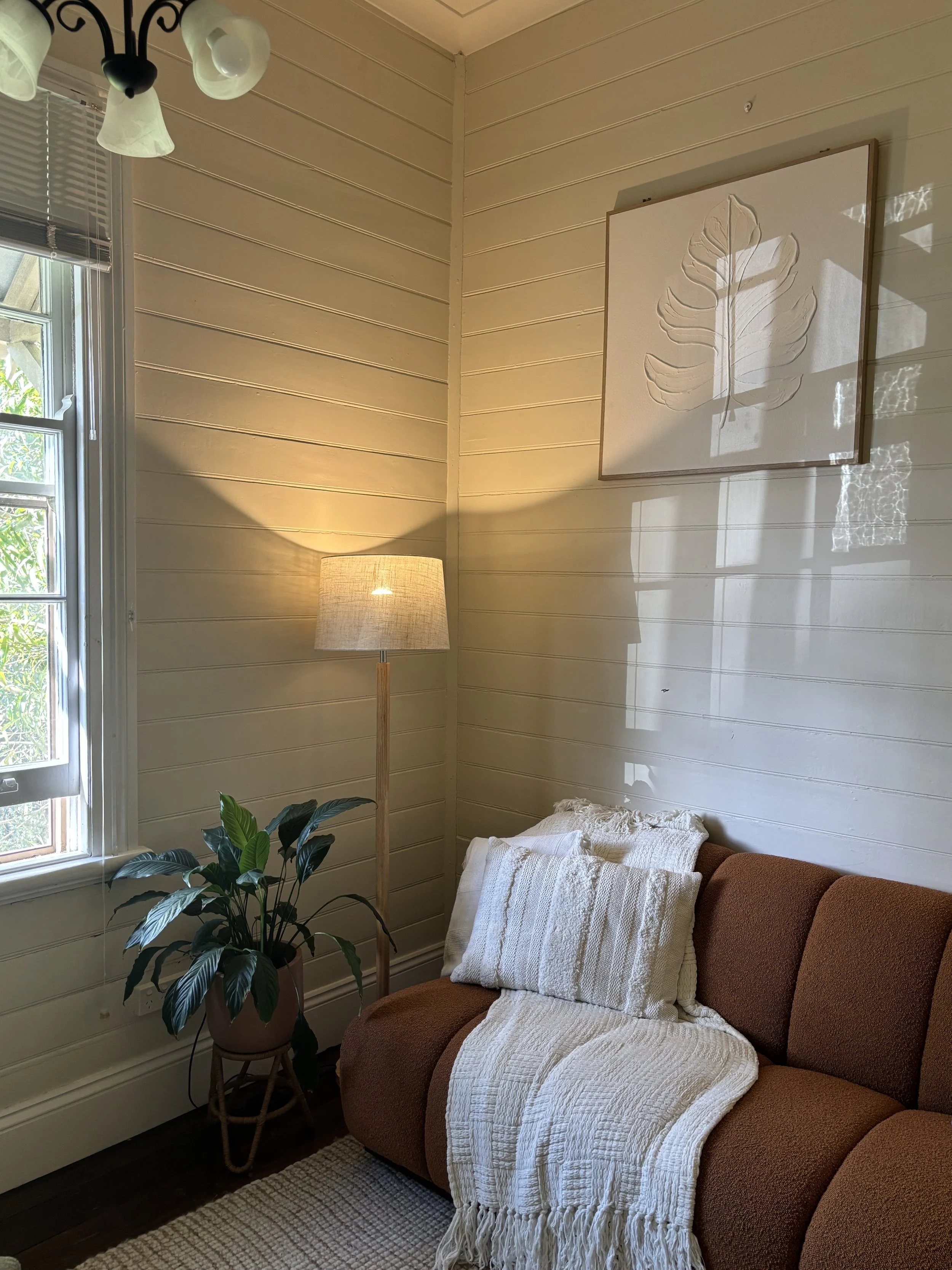 A cozy living room corner, a brown sofa draped with a cream knit blanket, a potted green plant on a small wooden stand, a tall floor lamp with a beige shade, and a wall art piece.  The Psych Nook, northern rivers Psychology practice.