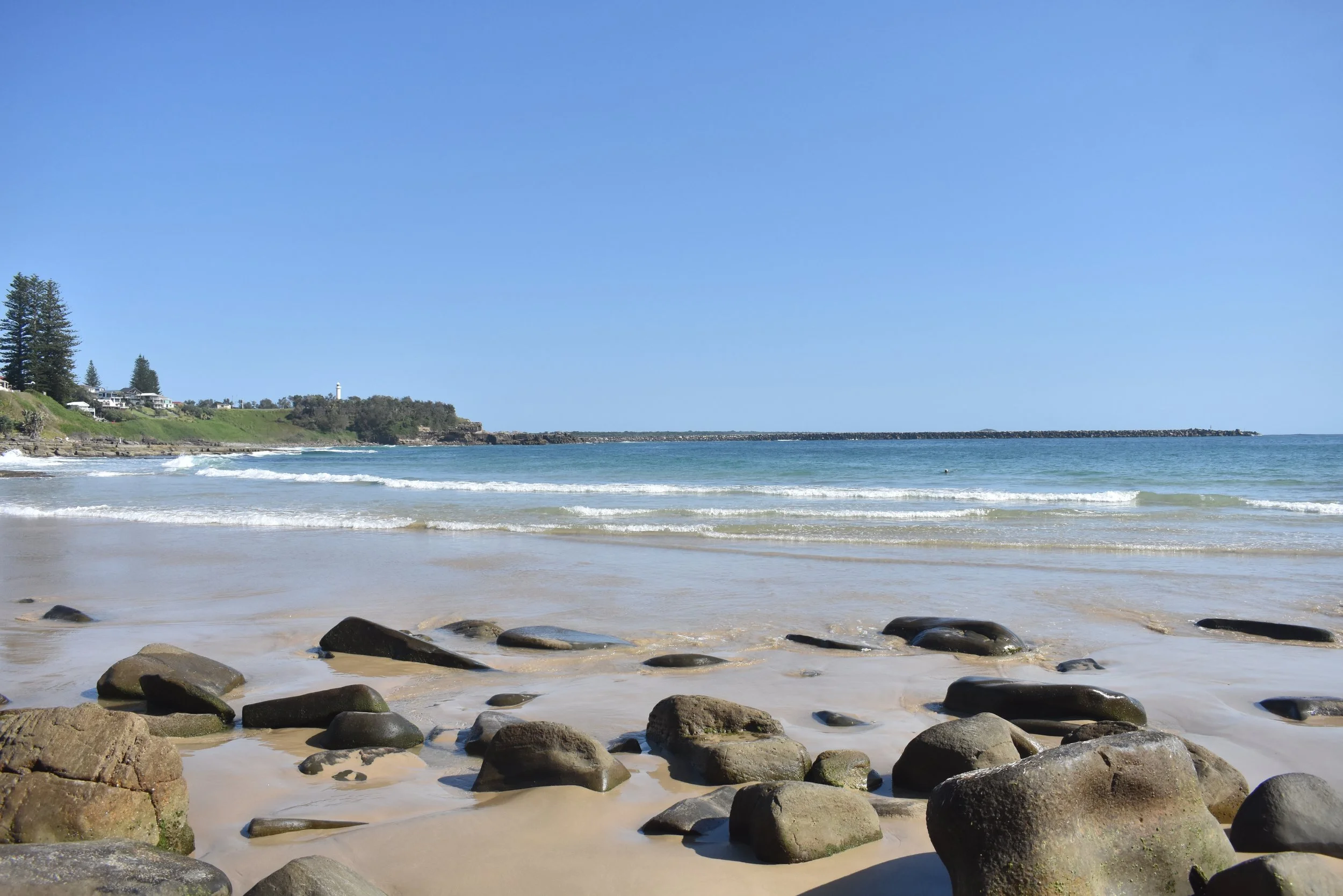 A scenic view of a beach with large rocks in the foreground, gentle waves, green cliffs with houses, and a lighthouse in the distance under a clear blue sky. Yamba, a small coastal town in the clarence valley.