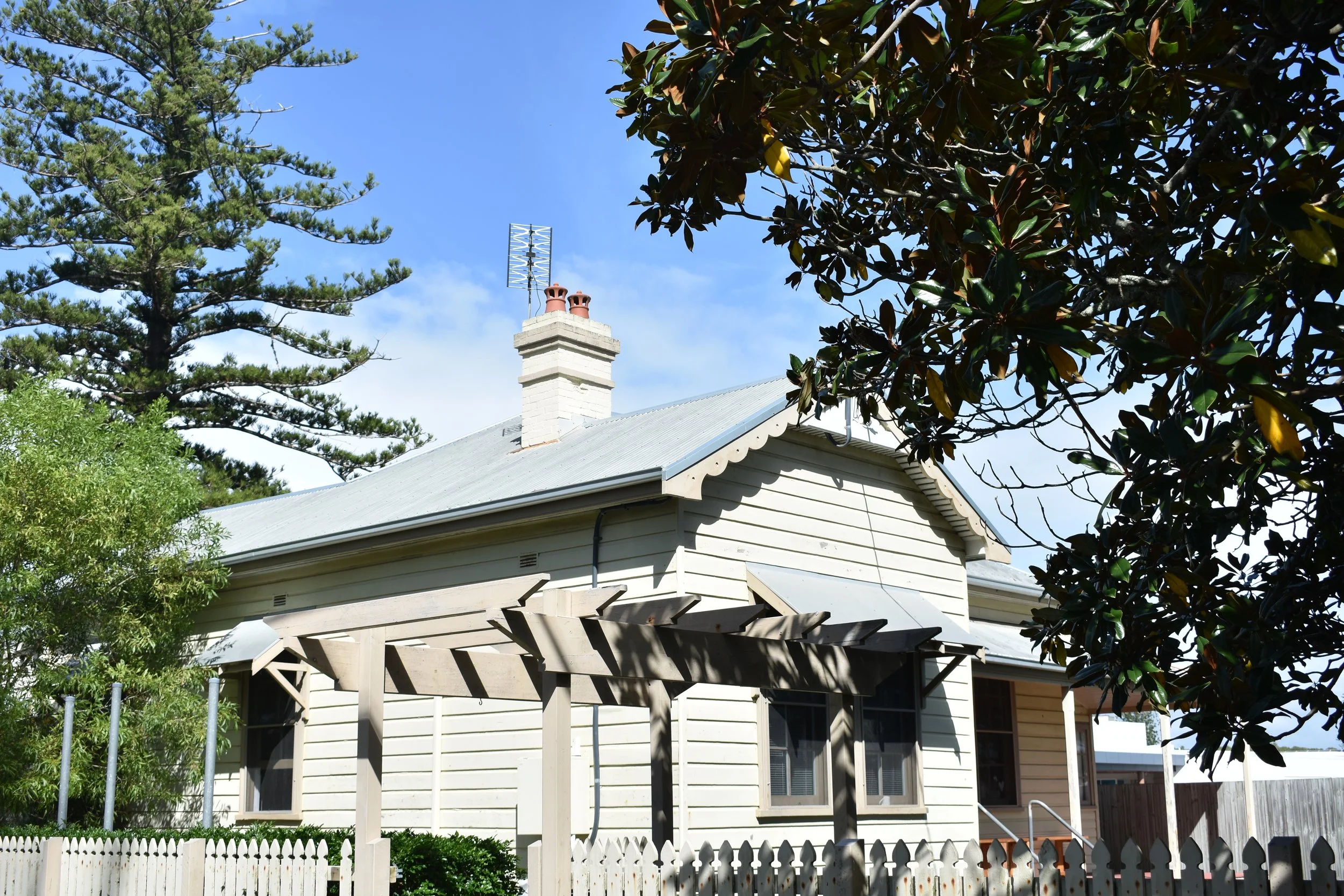 A house with a gable roof and a white picket fence in front, surrounded by trees, including pine and magnolia, under a clear blue sky.