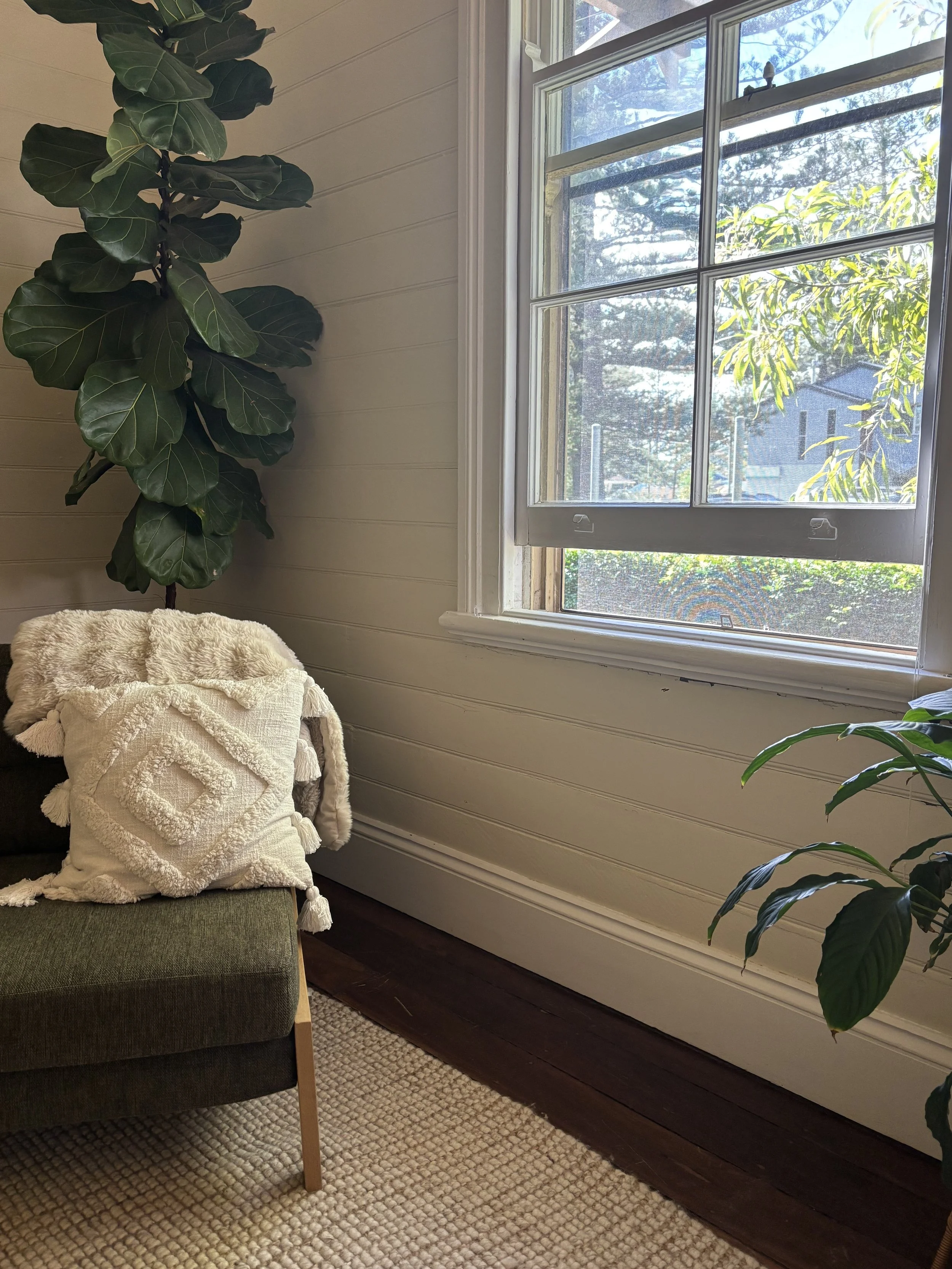A cozy corner of a room featuring a dark green armchair with a white textured throw blanket and two white pillows, beside a large houseplant with broad, dark green leaves. There is a window with a white frame that lets in natural light, and outside trees and part of a house are visible. The room has white-painted wooden walls and a beige woven rug on a dark wood floor.