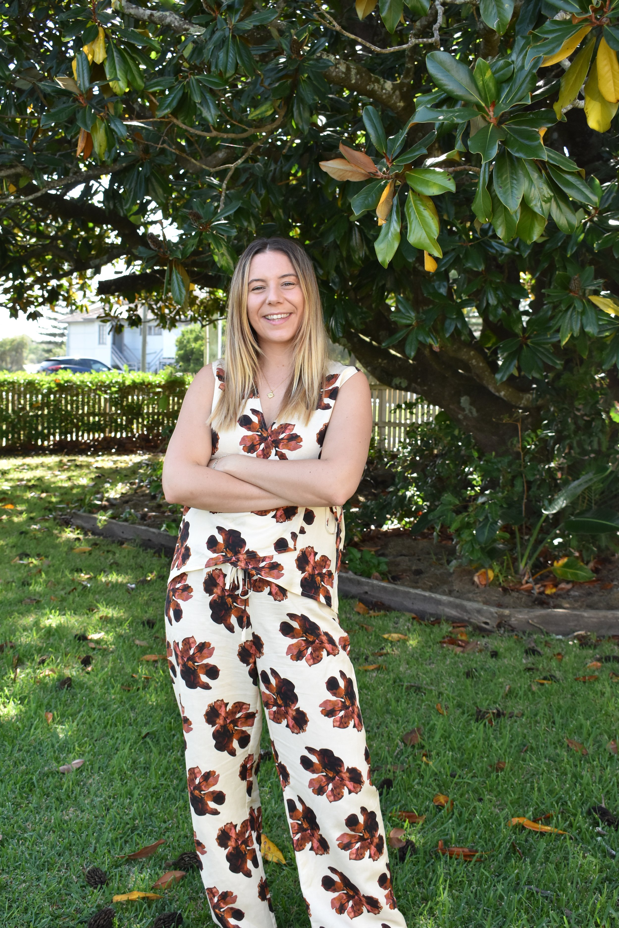 A young woman with blonde hair, smiling and with arms crossed, standing outdoors in front of a large leafy tree. She is wearing a sleeveless, cream-colored jumpsuit with a brown floral pattern.