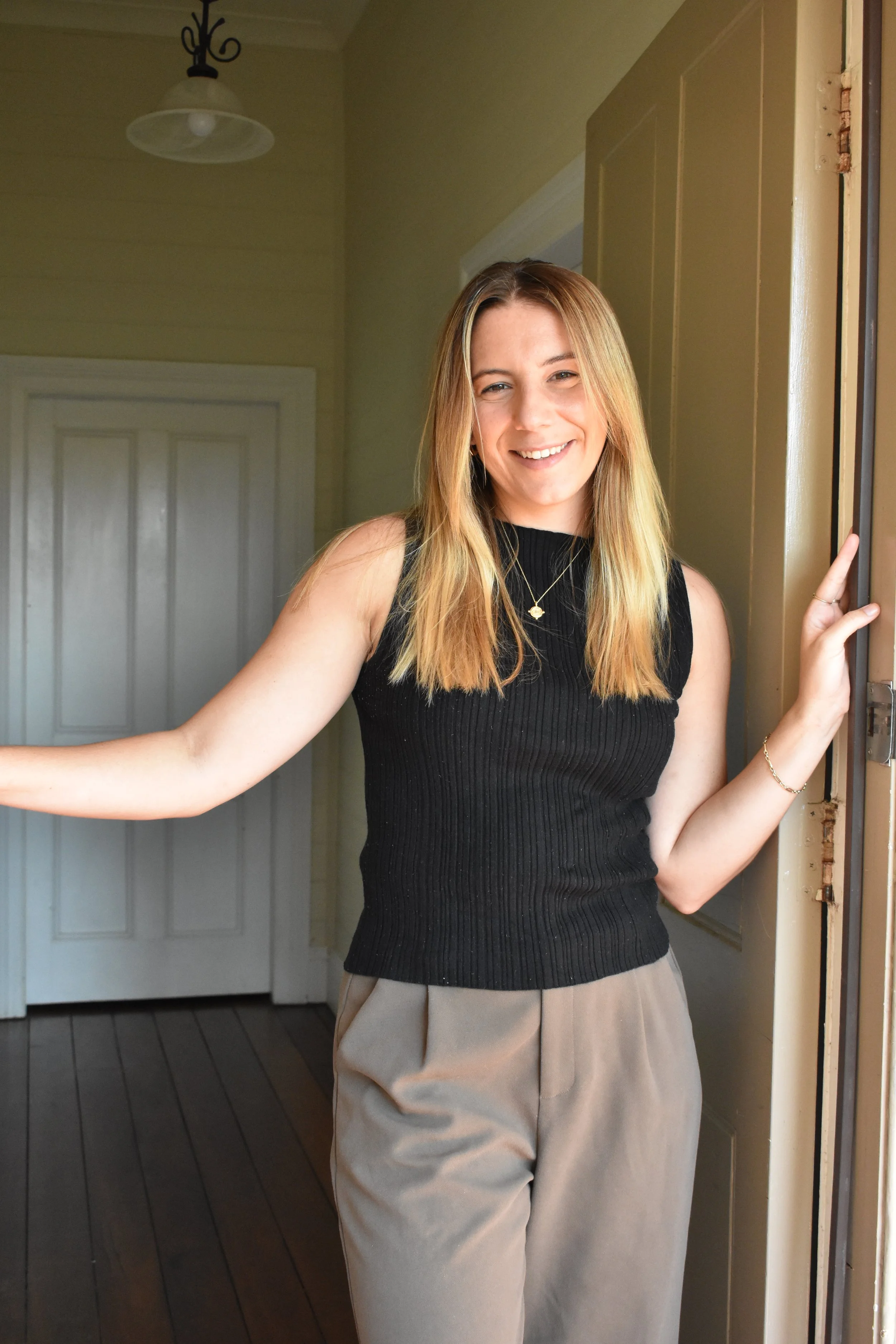 A young woman with long, blonde hair wearing a black sleeveless top and beige pants, smiling and standing next to a doorway in a cozy room with greenish walls and wooden flooring.