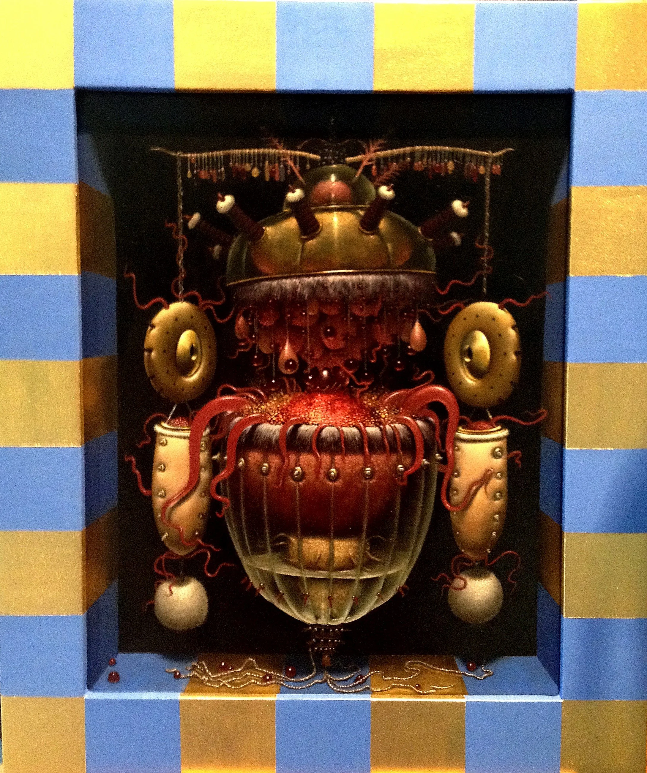 A decorative display of a mechanical or ornate mask or sculpture with orange, gold, and dark colors, featuring circular and red wire-like elements, surrounded by a blue and gold striped boxed frame.