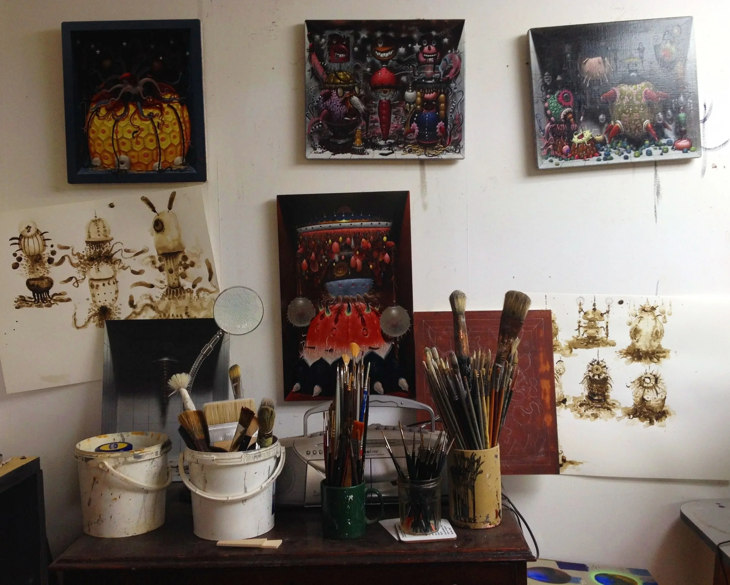Art studio workspace with paintings and art supplies, including buckets filled with brushes, some with dried paint, and paintings on the wall depicting colorful and surreal characters and scenes.