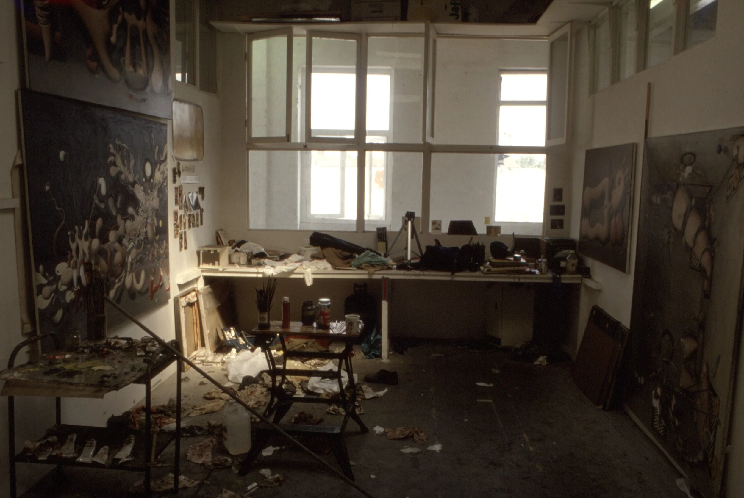 A cluttered art studio with paintings, tables, art supplies, and large windows letting in natural light, with some debris and mess on the floor.