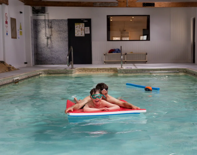 Two children on a red pool float in an indoor swimming pool, wearing goggles, with a boy behind and a girl in front, swimming together.