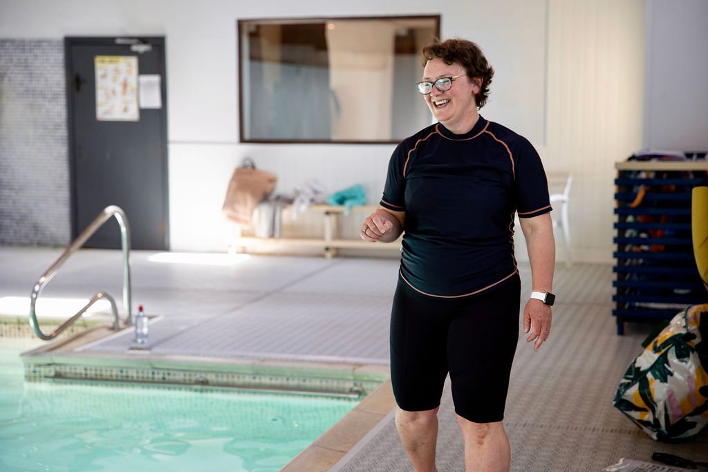 A woman wearing glasses and athletic clothing, smiling and walking near an indoor swimming pool.