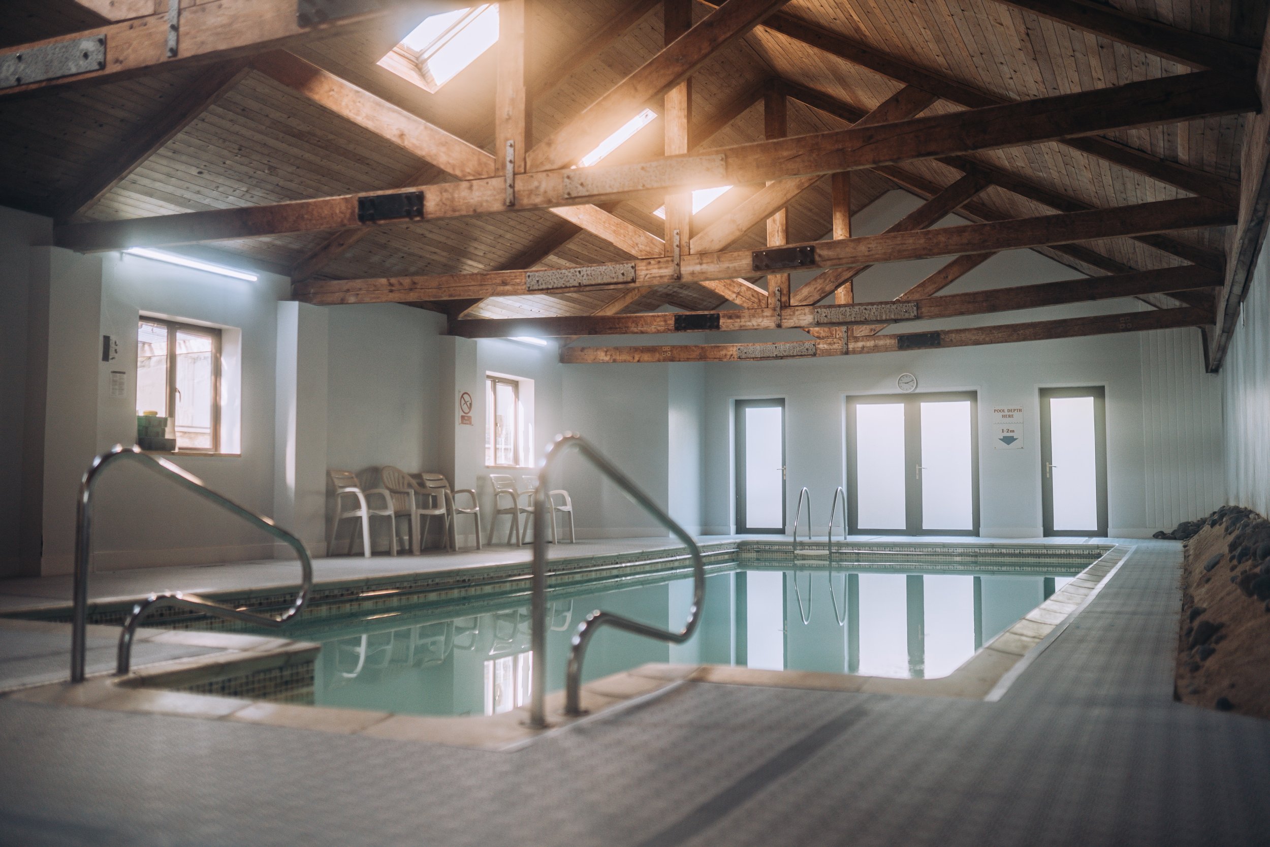Indoor swimming pool with wooden ceiling beams, multiple windows, and a set of chairs along the wall.