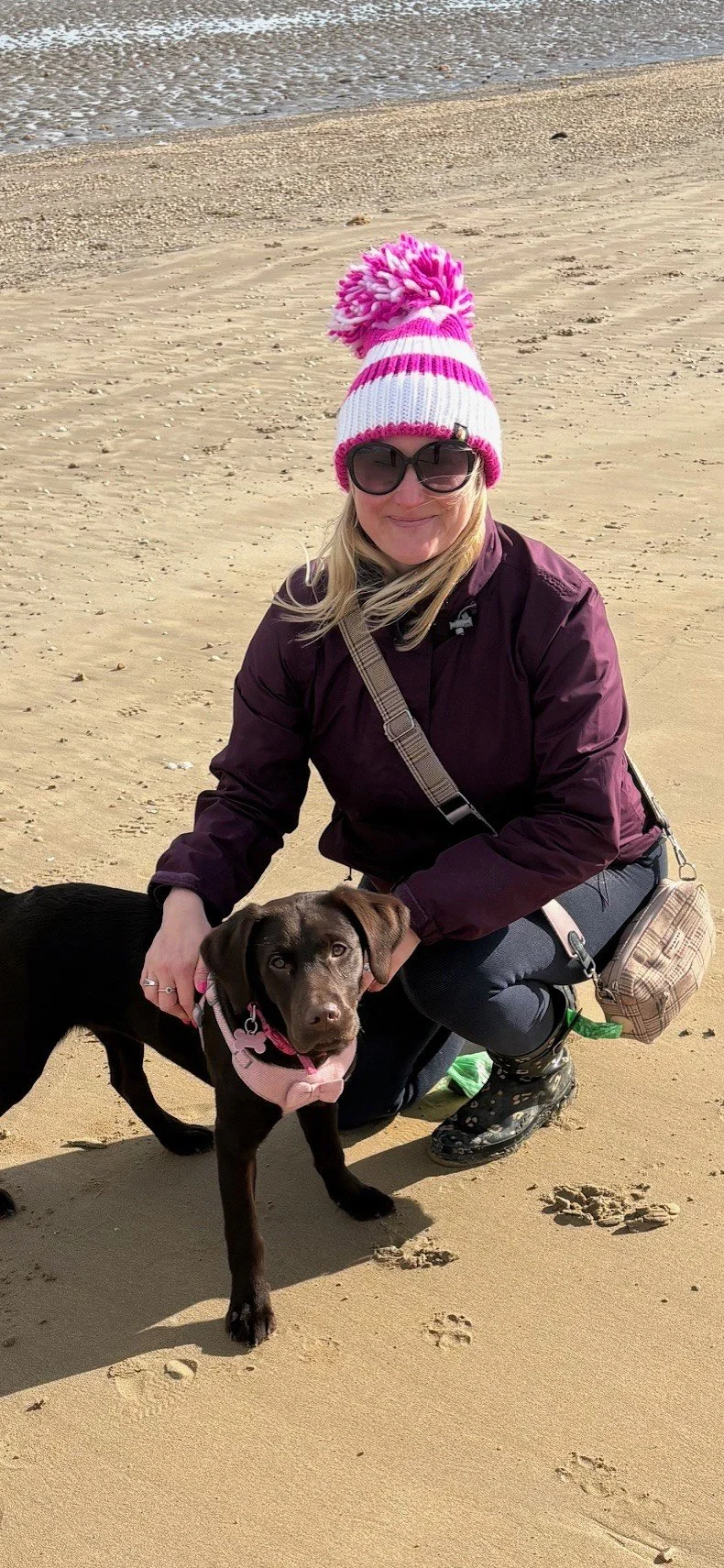 A woman in sunglasses and a pink striped knit hat with a pom-pom, kneeling on a sandy beach beside a chocolate Labrador puppy wearing a pink harness.