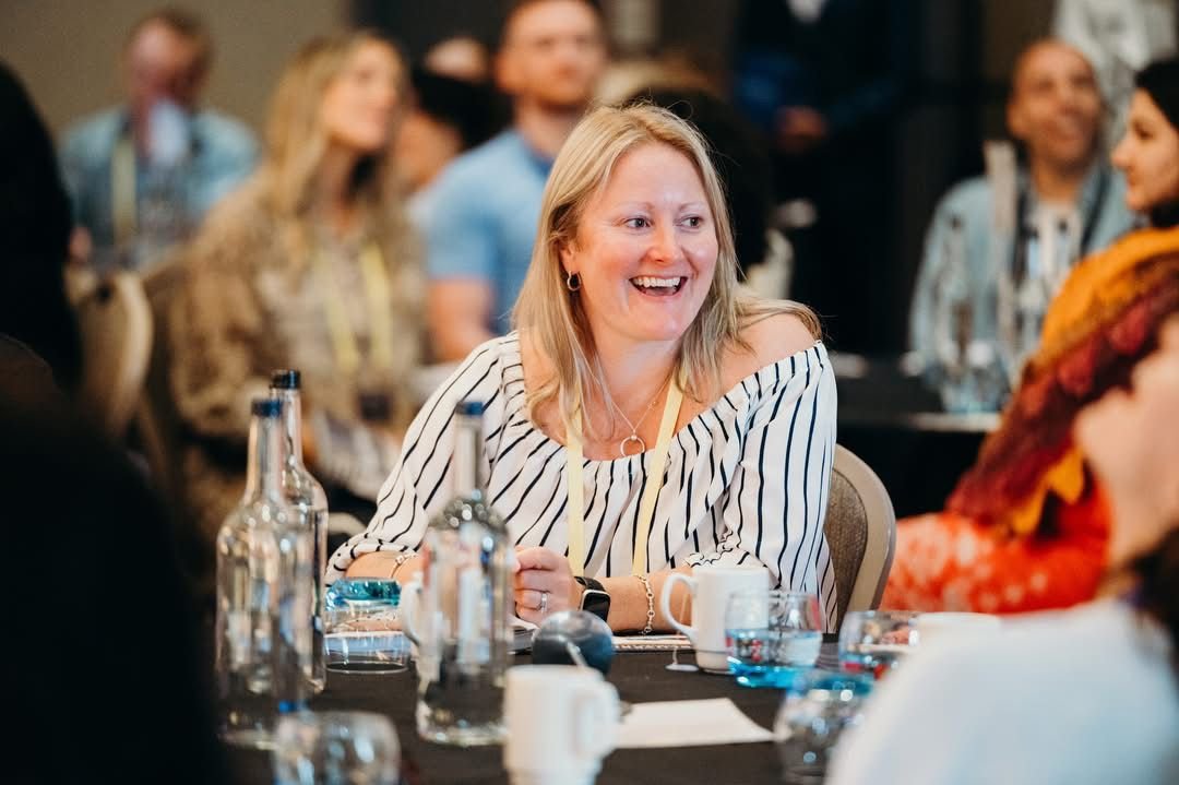 A blonde woman in a striped shirt smiling at a conference or meeting, sitting at a table with bottles, mugs, and glasses, surrounded by other people in a social or professional setting.