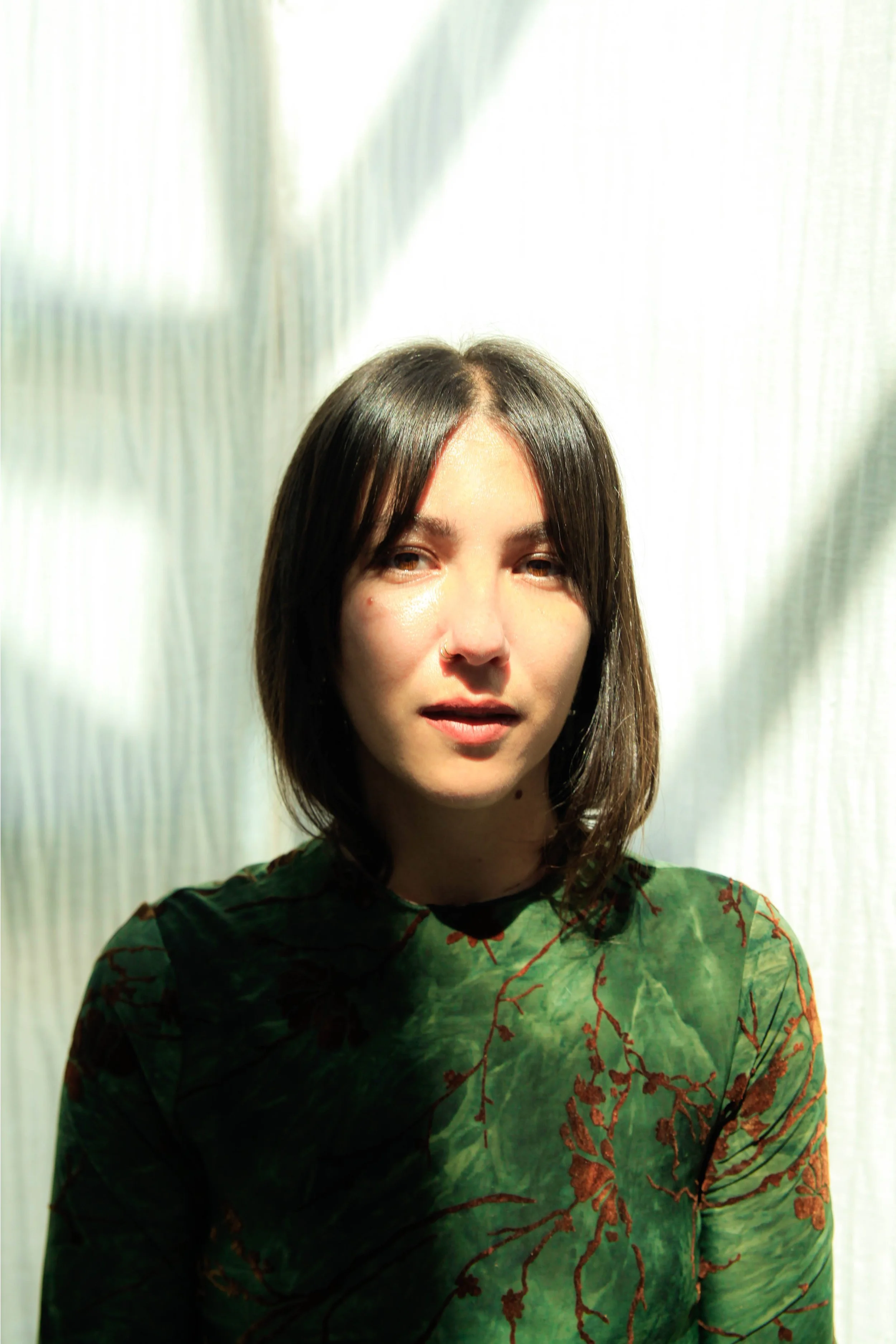 Portrait of a young woman with shoulder-length dark hair, wearing a green patterned shirt, standing in front of a plain light-colored curtain.