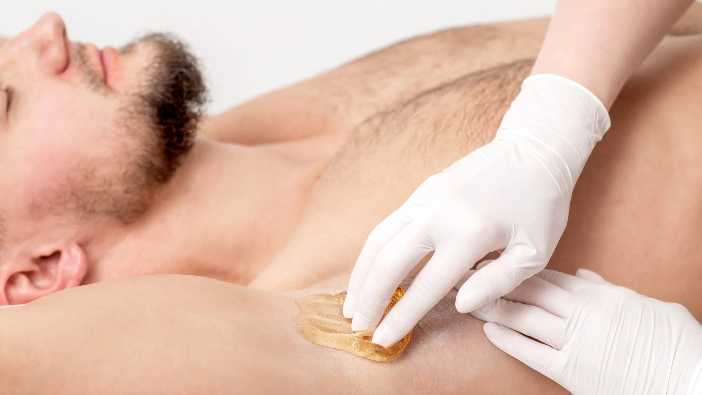 A man lying down with closed eyes and a beard, receiving a waxing treatment on his armpit from a person wearing white gloves.