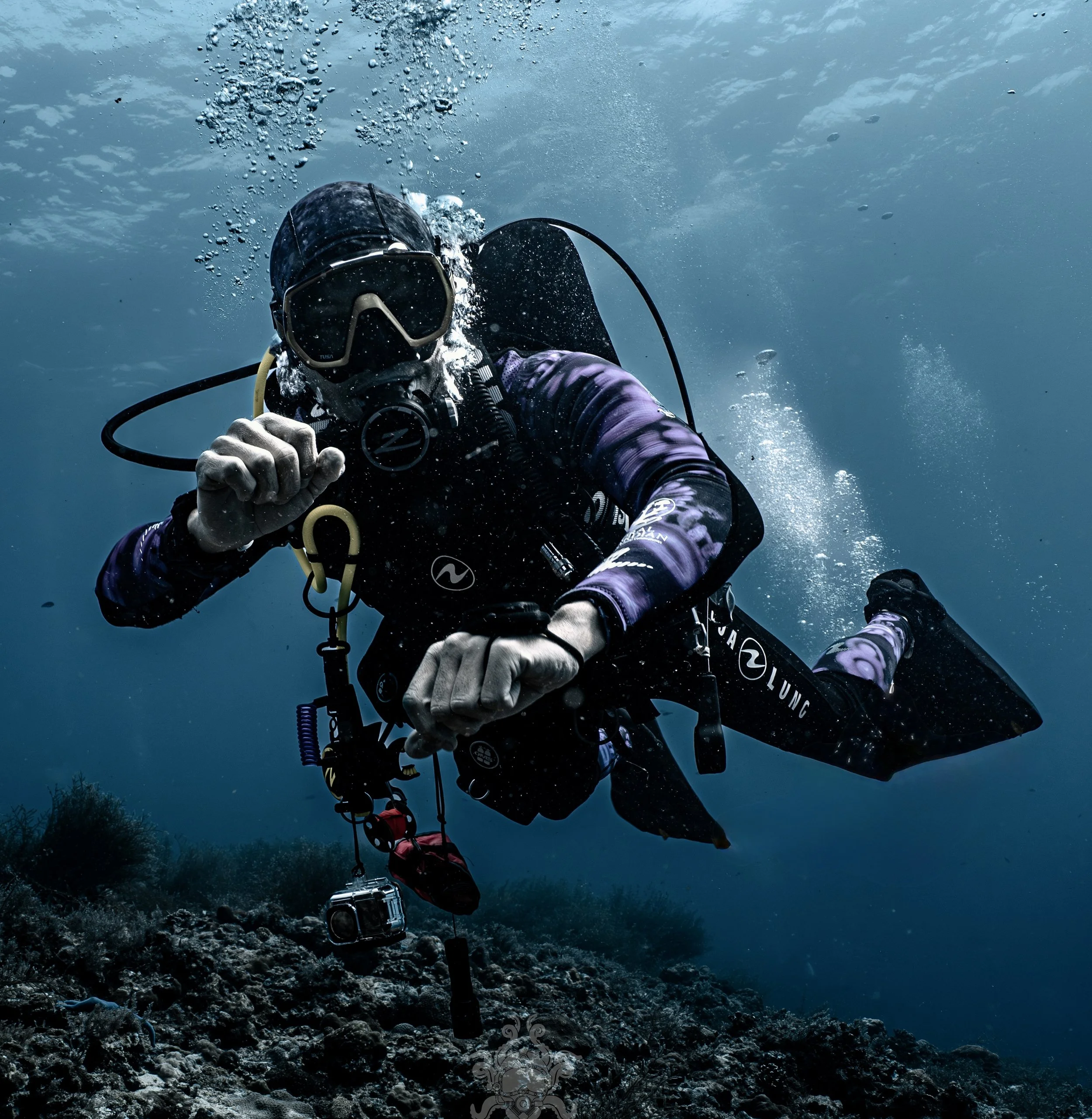 A scuba diver wearing a mask, wetsuit, and holding a camera, submerged underwater with particles of bubbles and water around, ascending towards the surface near a rocky bottom.