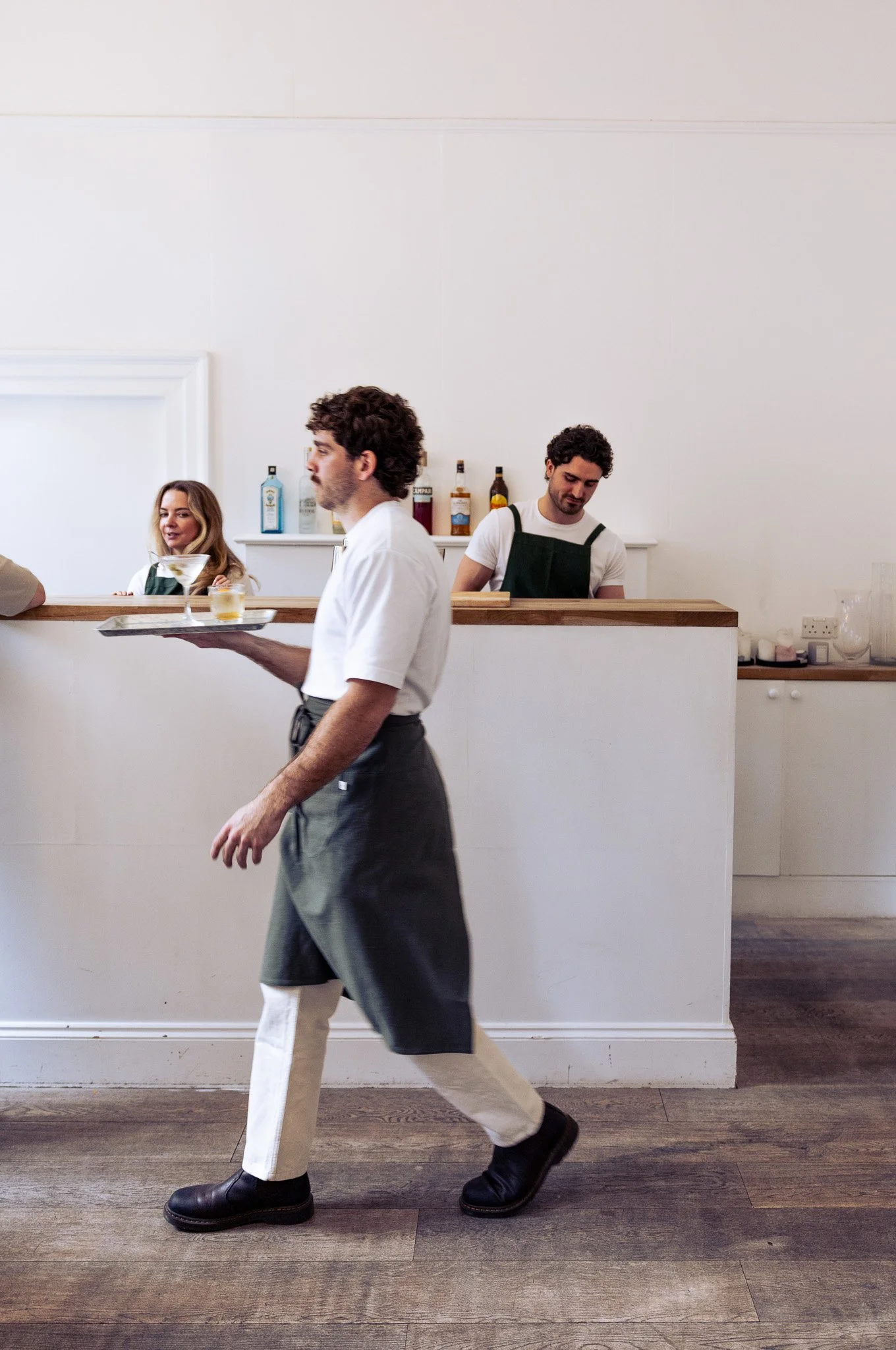 A waiter in a black apron and white shirt carrying a drink in a cafe or restaurant.