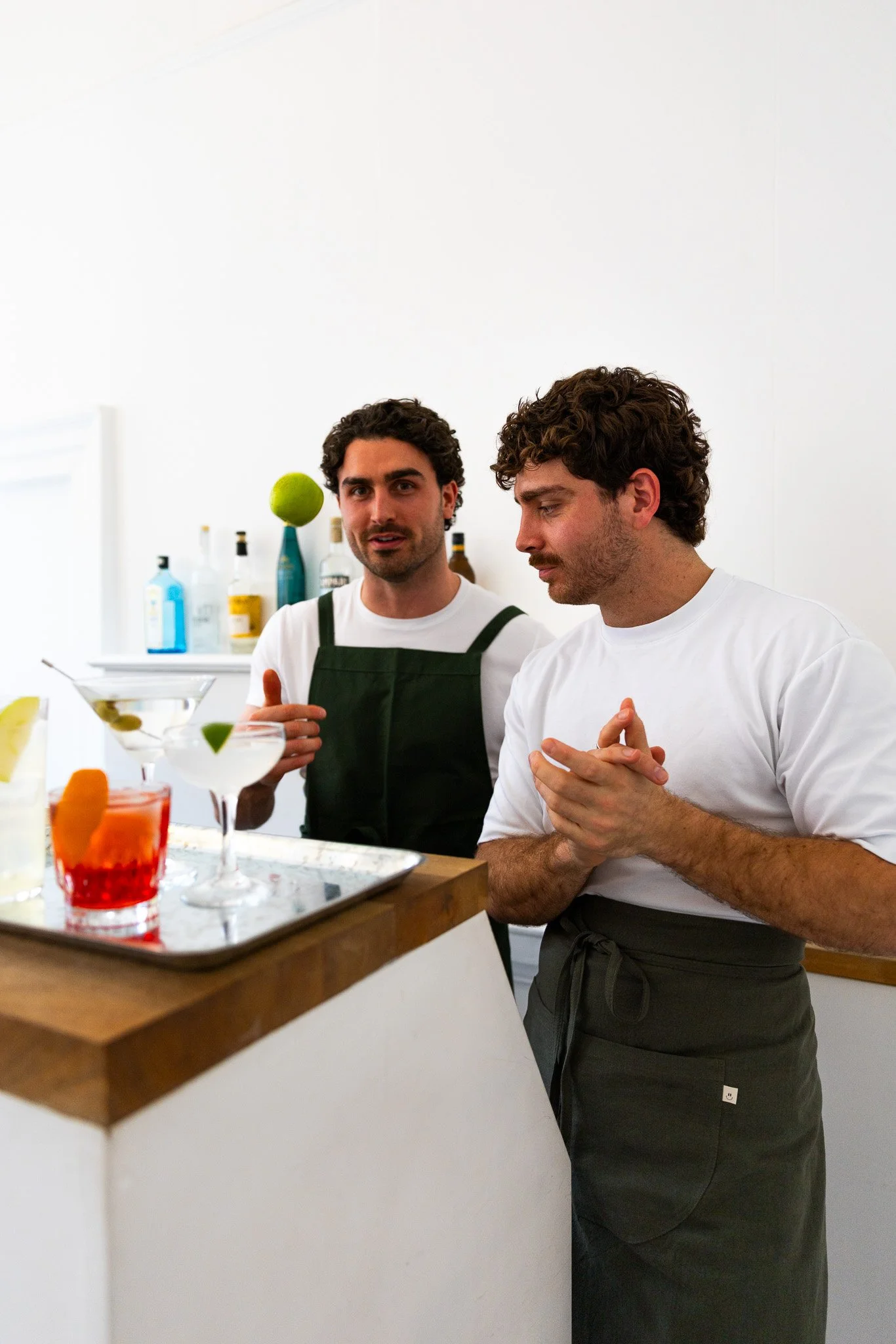 Two bartenders preparing colorful cocktails at a bar counter in a bright, minimalist setting.