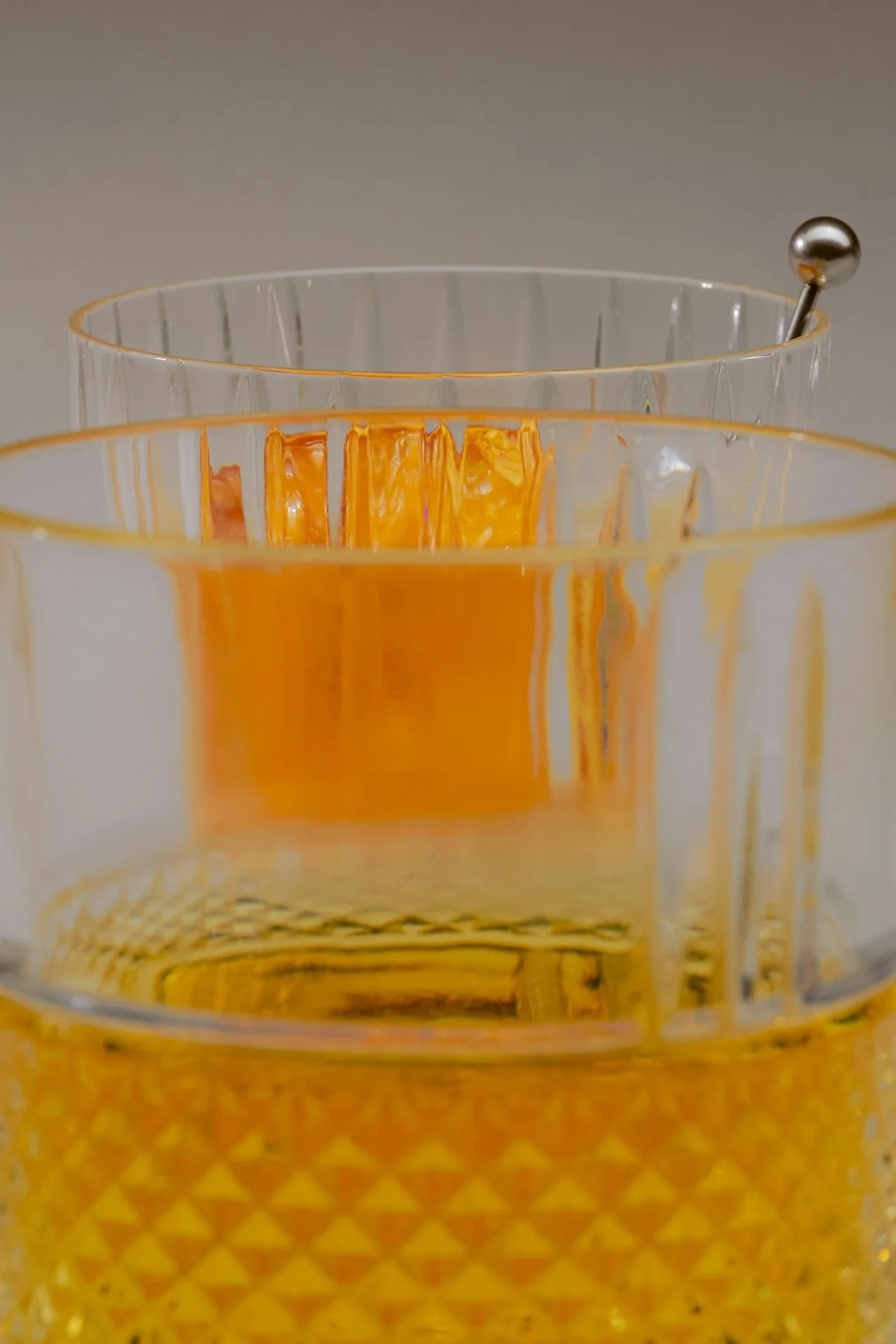 Close-up of two ornate glasses, one with orange liquid and a cocktail pick, against a neutral background.