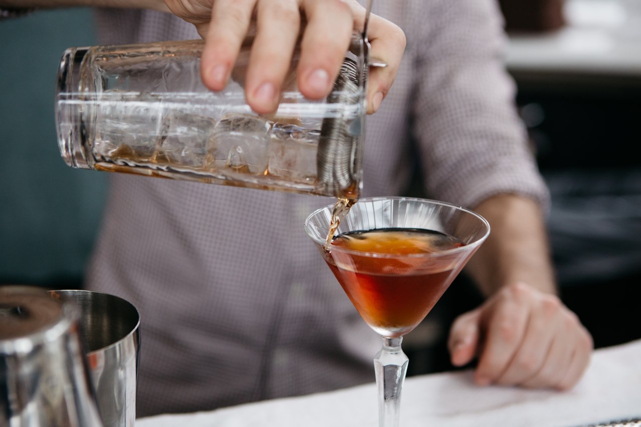 Person pouring a brown cocktail into a martini glass with a straw, ice in a glass container nearby.