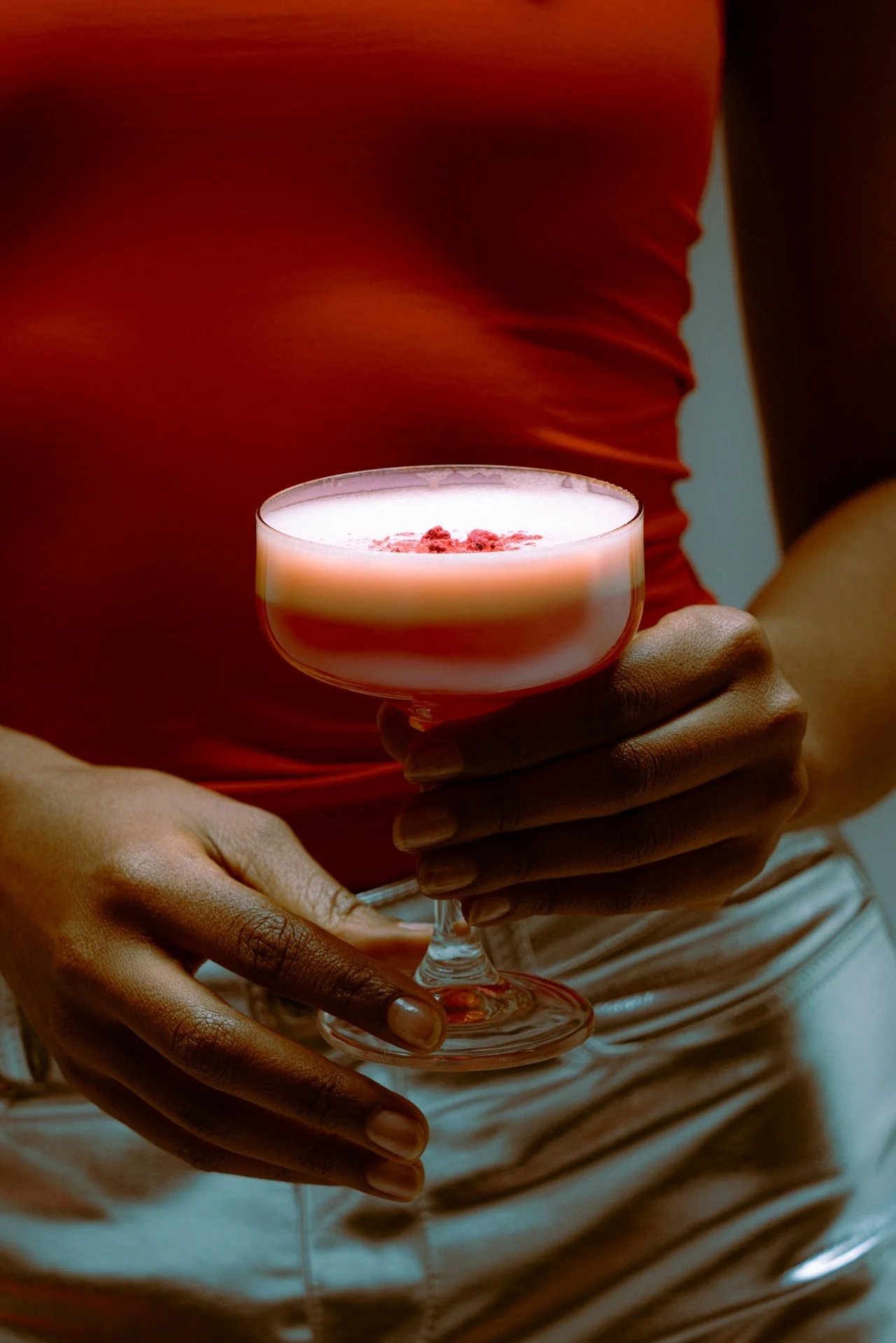 Person wearing a red shirt holding a cocktail glass with a pink-colored drink garnished with raspberries.
