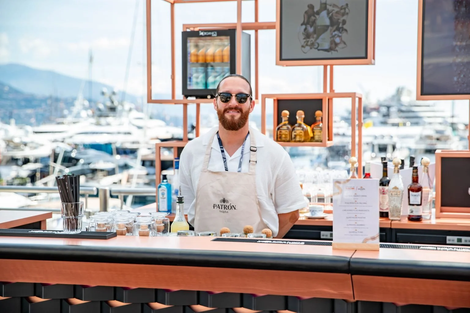 A smiling man with sunglasses, a beard, and a white shirt stands behind a bar at a marina, with boats and yachts in the background. He wears an apron with the Patrón logo.