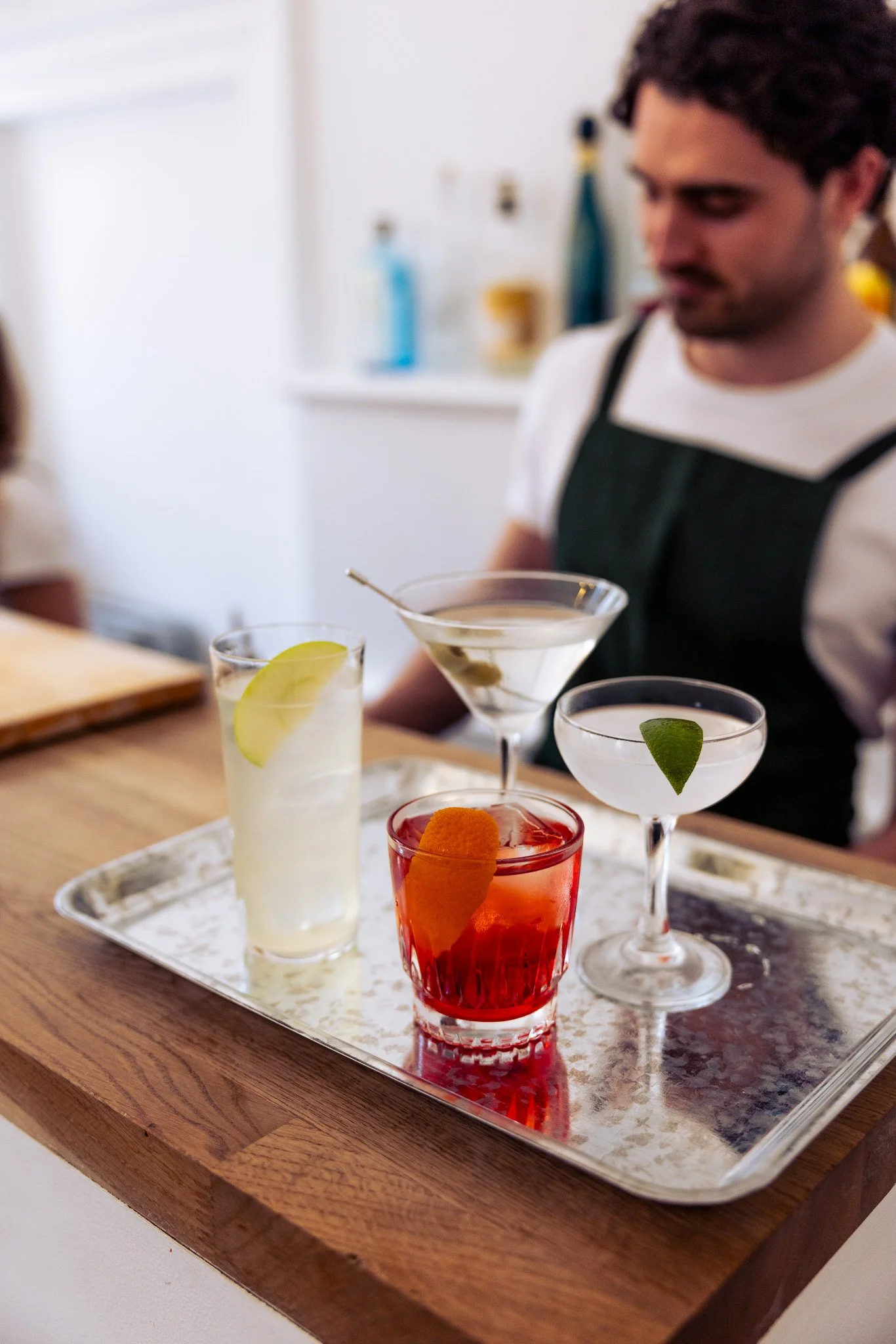 A silver tray with four colorful cocktails on a wooden table, with a bartender in the background.