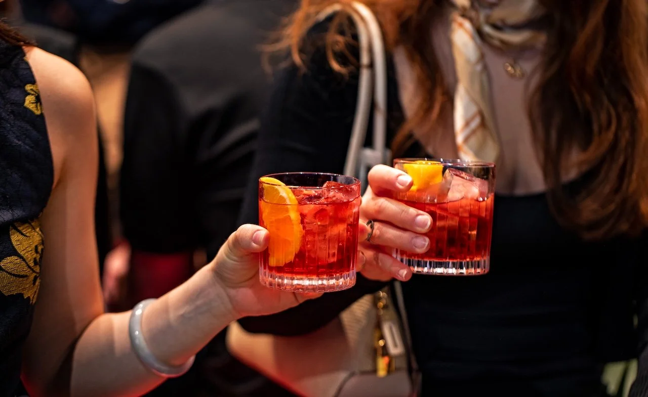 Two women holding glasses of red-colored cocktails with orange slices at a social gathering.
