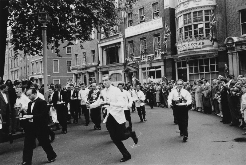 Black and white photo of a street parade with waiters carrying trays, crowds watching, and buildings with signs in the background.