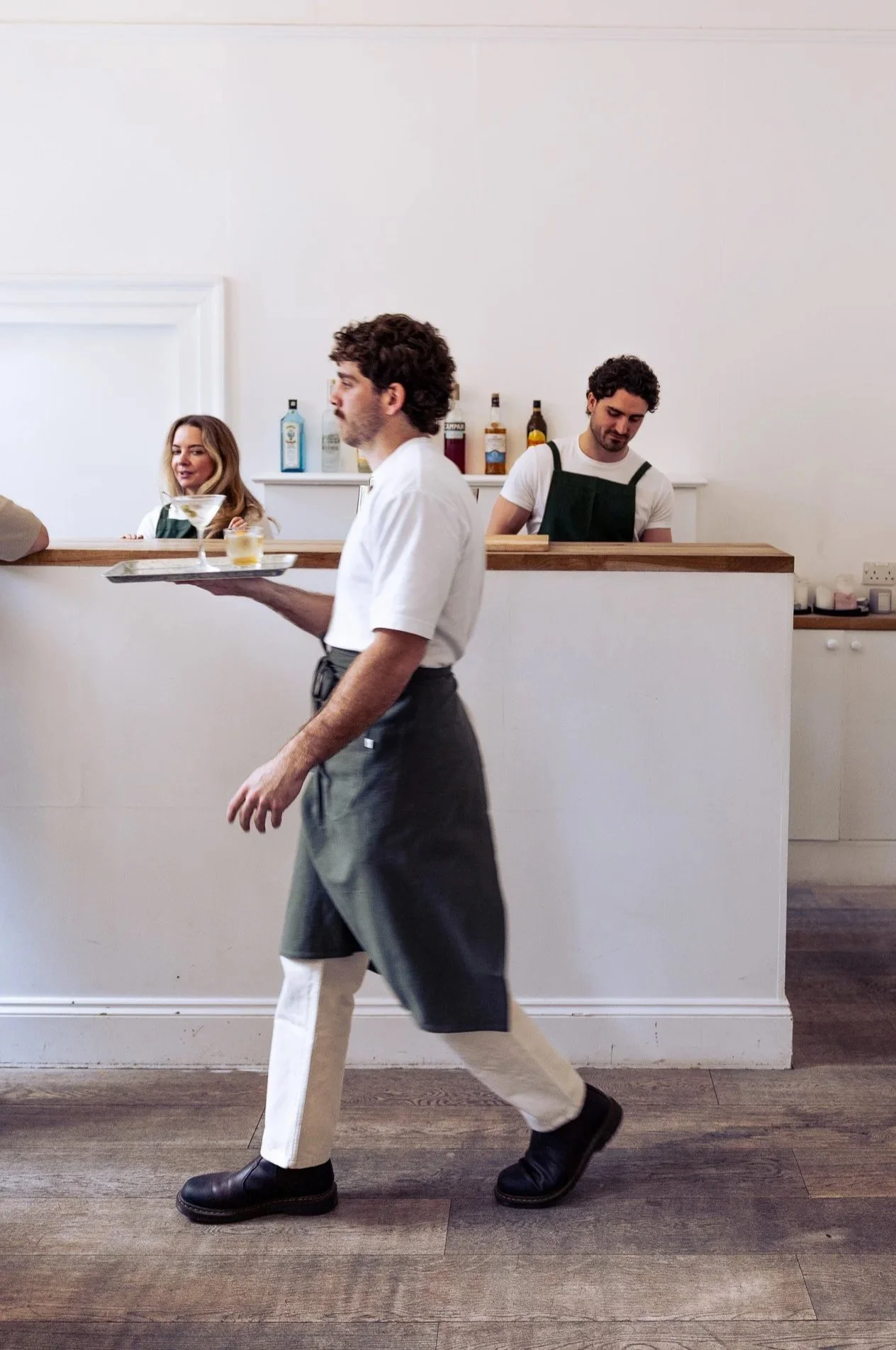 A bartender walking past the bar with drinks while two bartenders work behind the bar in a restaurant or bar setting.