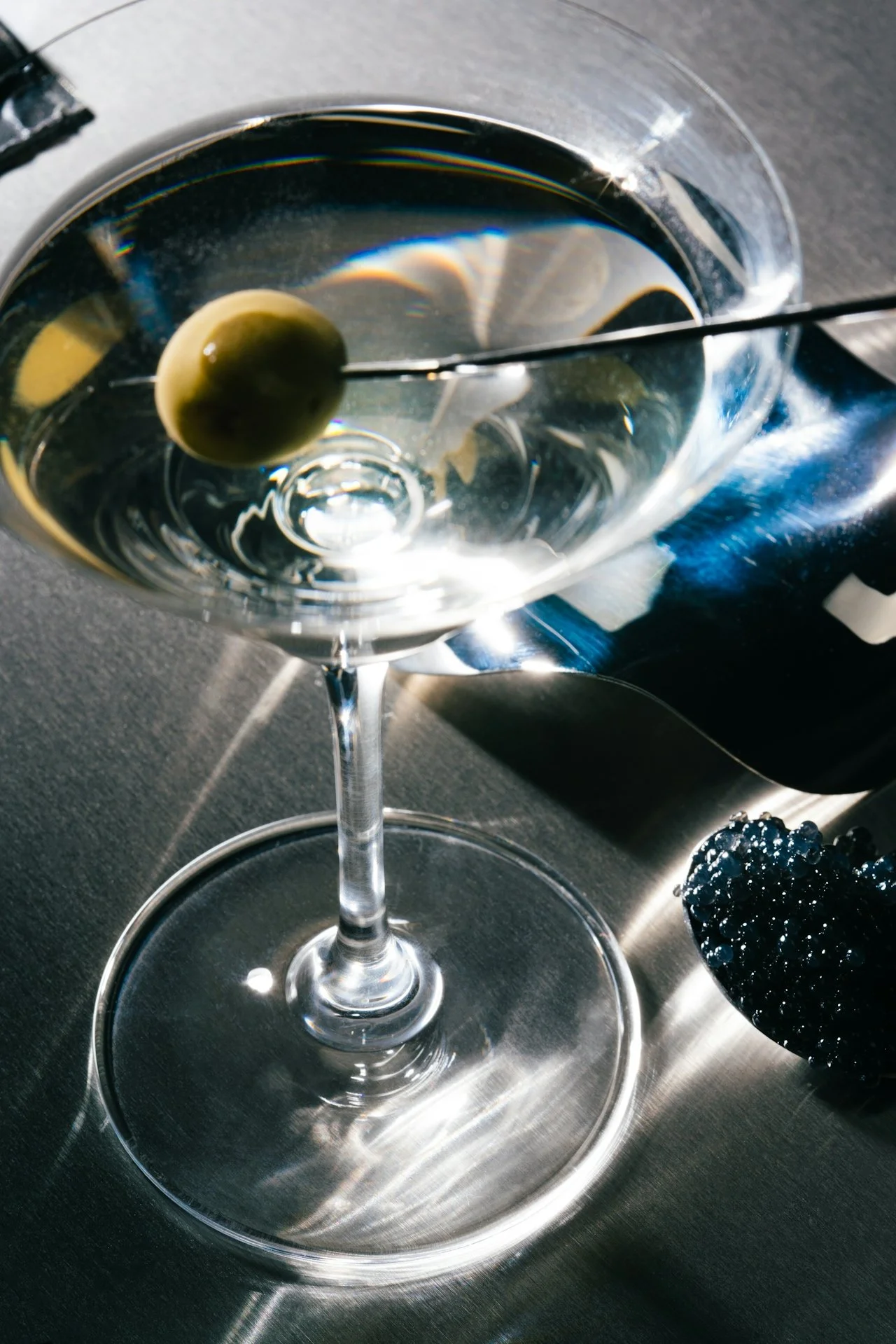 A martini glass with a clear liquid, garnished with an olive on a pick, on a metallic surface with sunlight reflections.