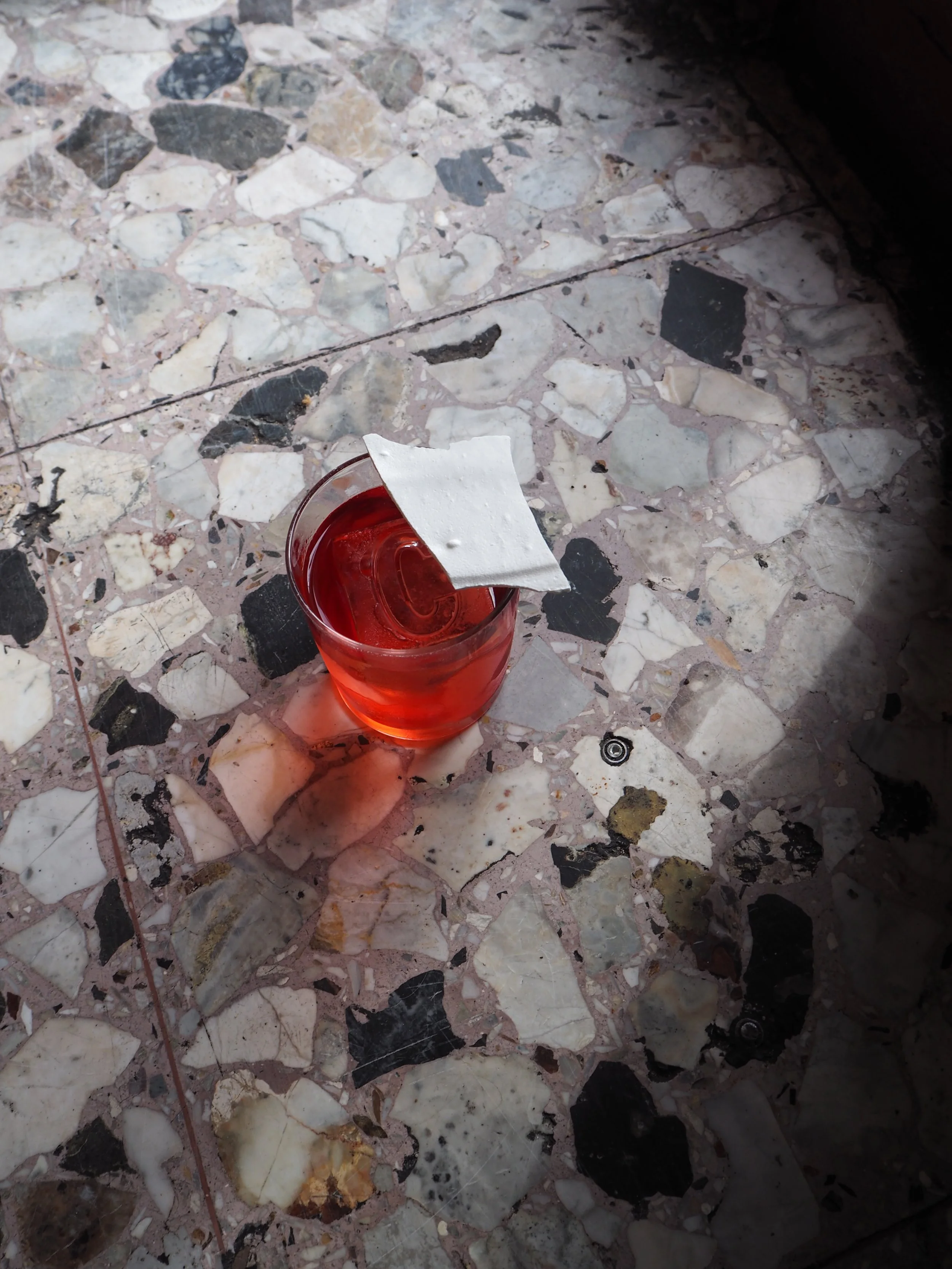 A glass of red beverage with a wet wipe resting on the rim, placed on a terrazzo floor.