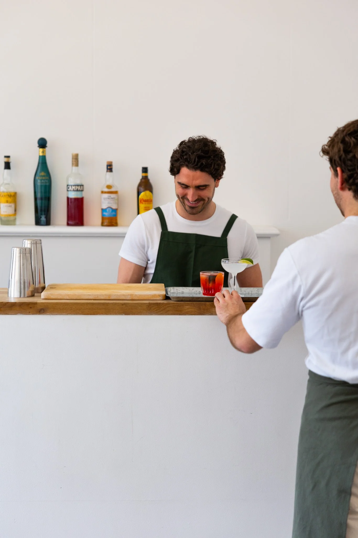 Bartender smiling at customer while serving drinks at a bar, with liquor bottles on shelf in the background.