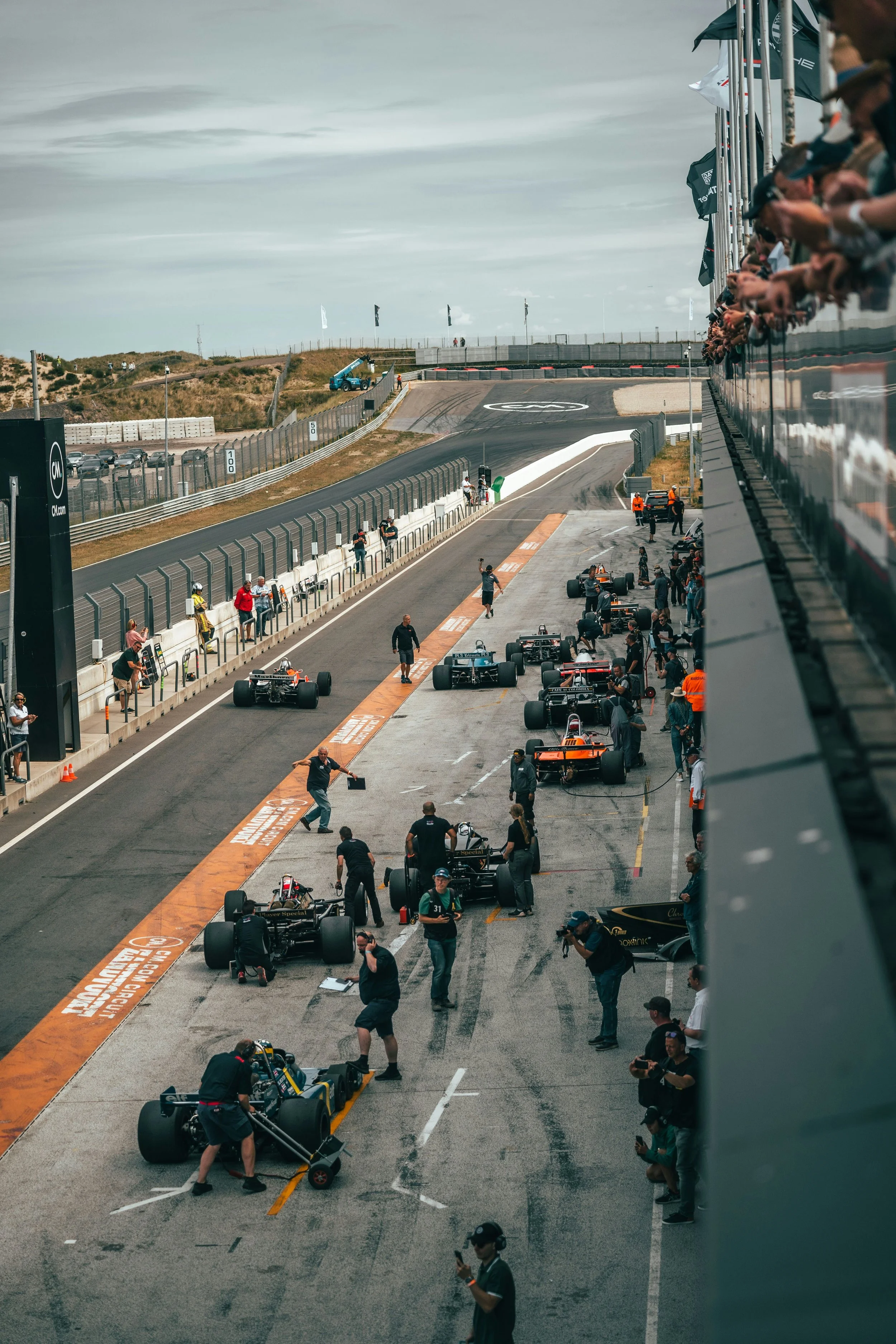 View of a Formula 1 race car pit stop at a track with team members working on cars, spectators watching from the grandstand, and the race track extending into the distance under a cloudy sky.
