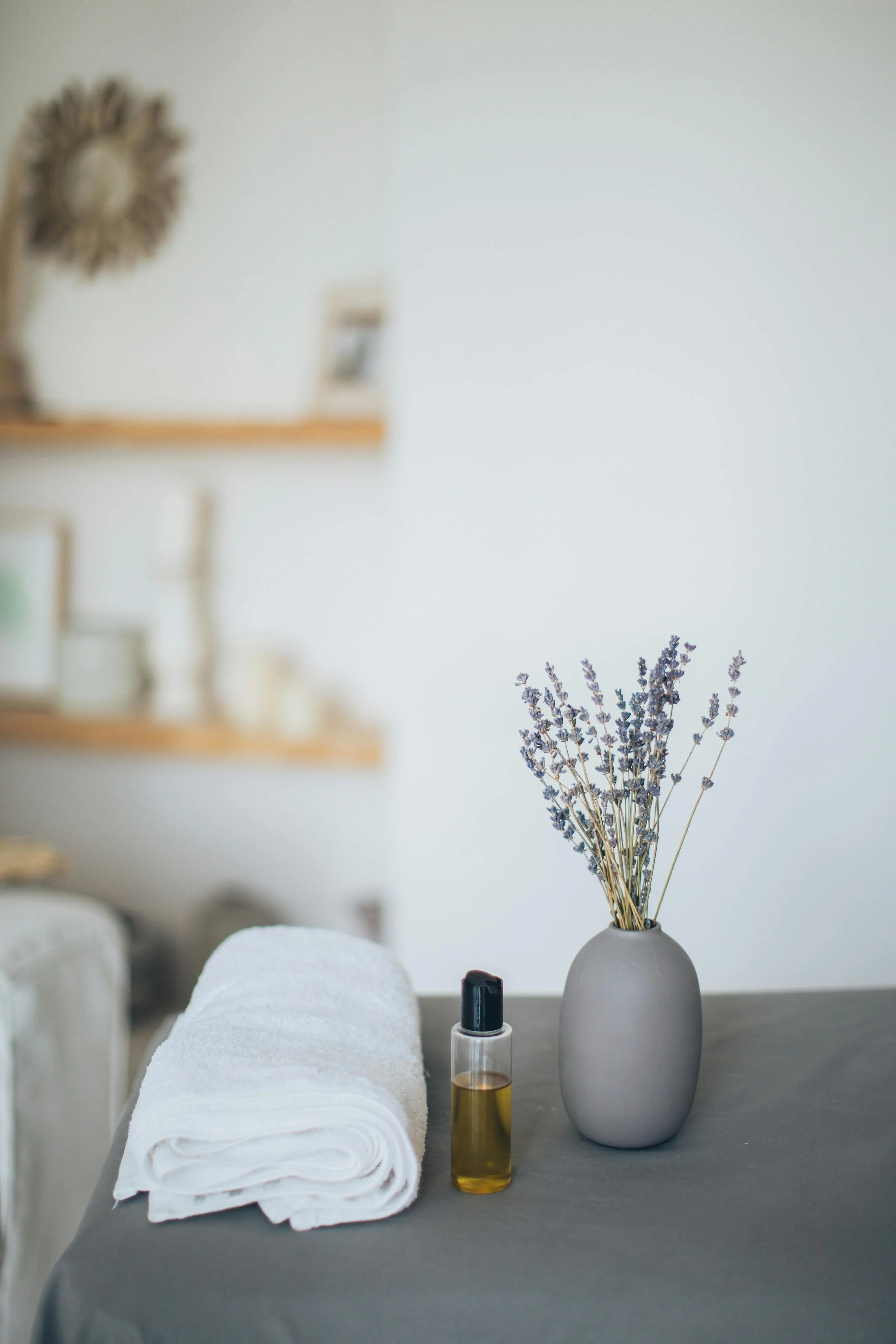 A rolled white towel, a small bottle of oil or lotion, and a vase with dried lavender are arranged on a gray tablecloth in a cozy, minimalist room with wooden shelves and wall decor in the background.