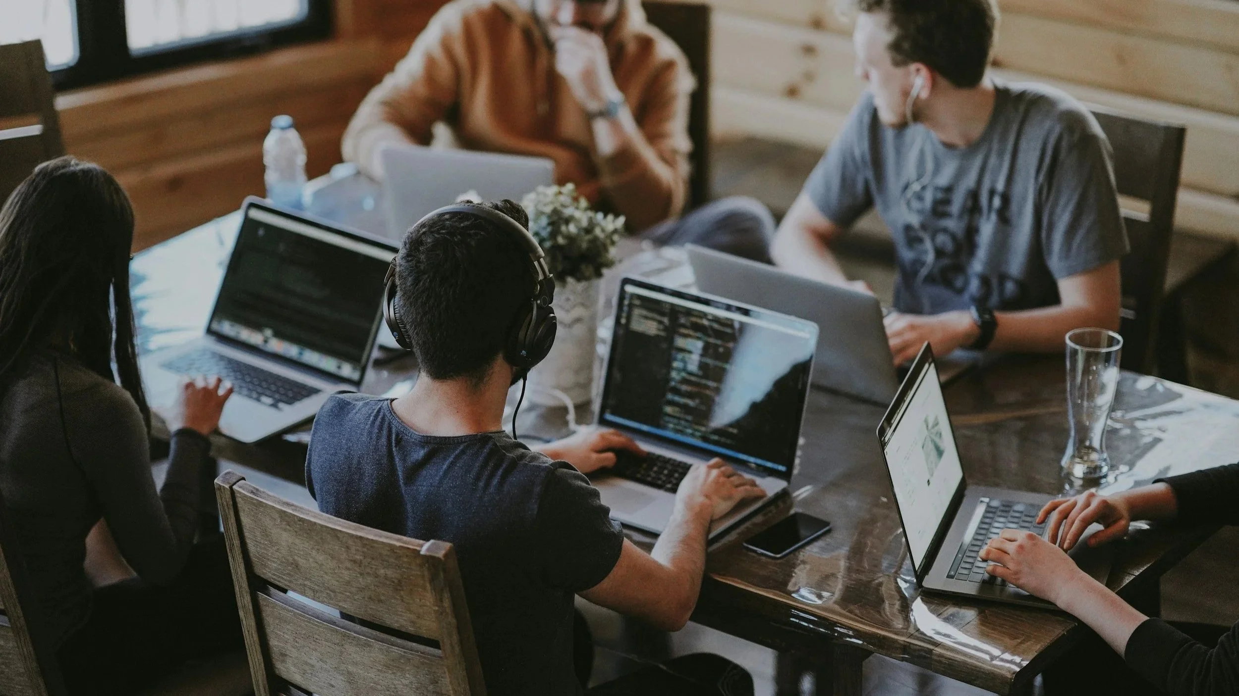 Menschen arbeiten an Laptops in einem modernen Raum, einige mit Kopfhörern, eine Person trägt ein graues T-Shirt, andere eine schwarze Jacke, es gibt Wasserflaschen und ein Smartphone auf dem Tisch.