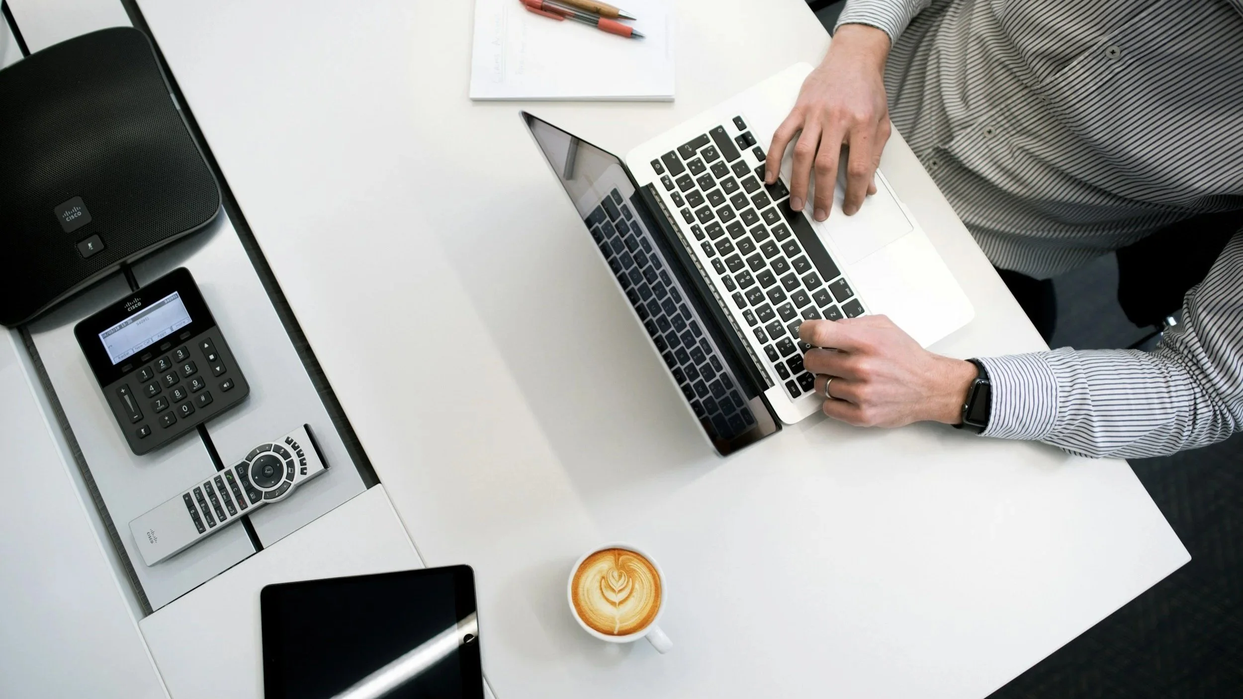 Ein Mann arbeitet an einem weißen Schreibtisch mit einem Laptop, mehreren Fernbedienungen, einem Smartphone, einem Notizbuch mit Stiften und einer Tasse Kaffee mit einem Herzverzierungen im Latte Art.
