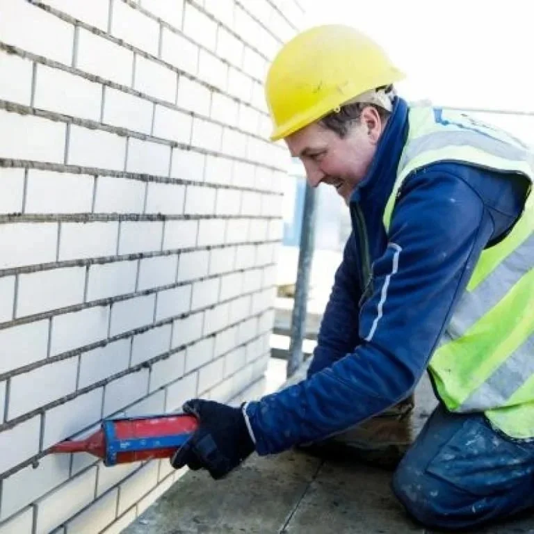 A construction worker wearing a yellow safety helmet, blue jacket, and reflective vest kneels on the ground, holding a red caulking gun, applying sealant to a brick wall.