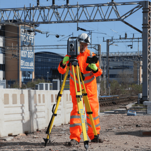 Construction worker wearing an orange safety suit, helmet, and gloves, operating a surveying instrument on a railway construction site with overhead electrical lines and industrial buildings in the background.