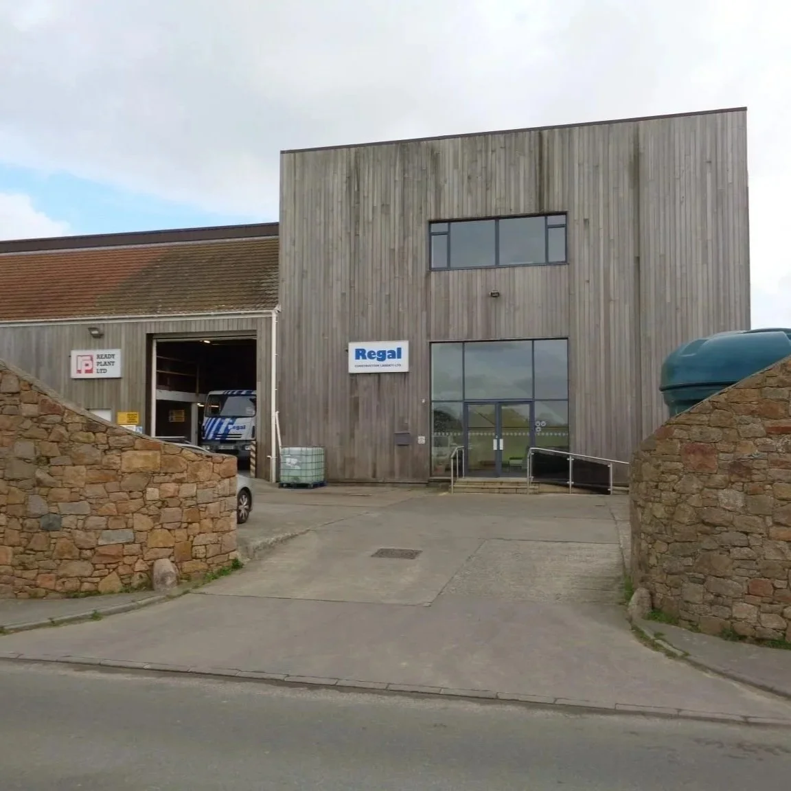 The image shows a commercial building with a wooden exterior, featuring large glass doors and windows. There is a sign on the building that reads 'Regal,' and a partially open garage door with a pickup truck inside. In the foreground, there are stone walls and a sloped driveway leading to the entrance.