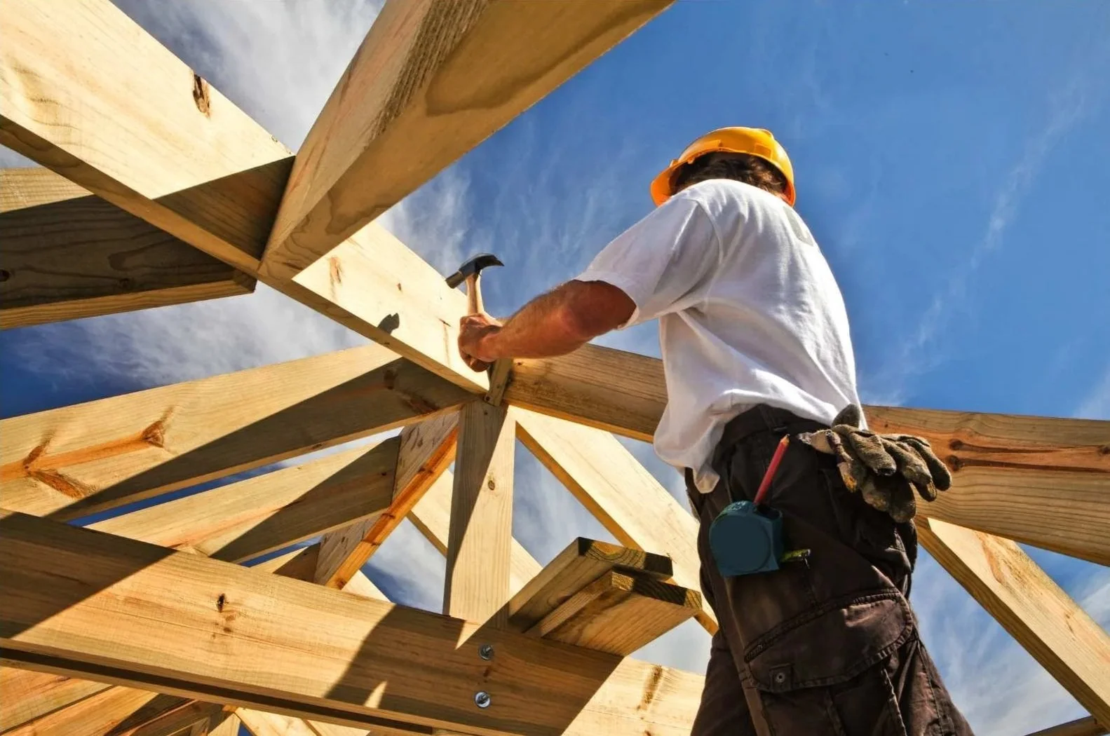 A construction worker wearing a yellow helmet and white t-shirt hammering a nail into a wooden beam during construction of a wooden structure. The sky is visible in the background.