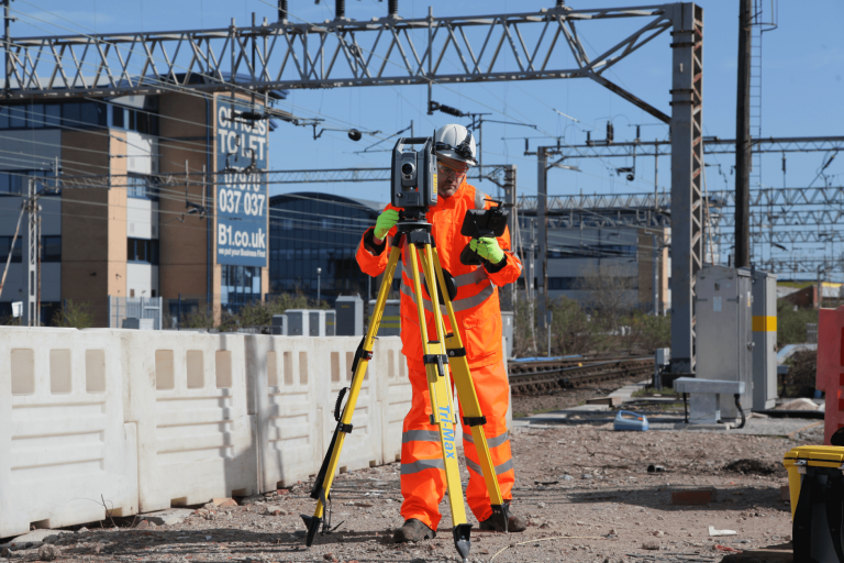 A worker in safety gear using surveying equipment next to train tracks.
